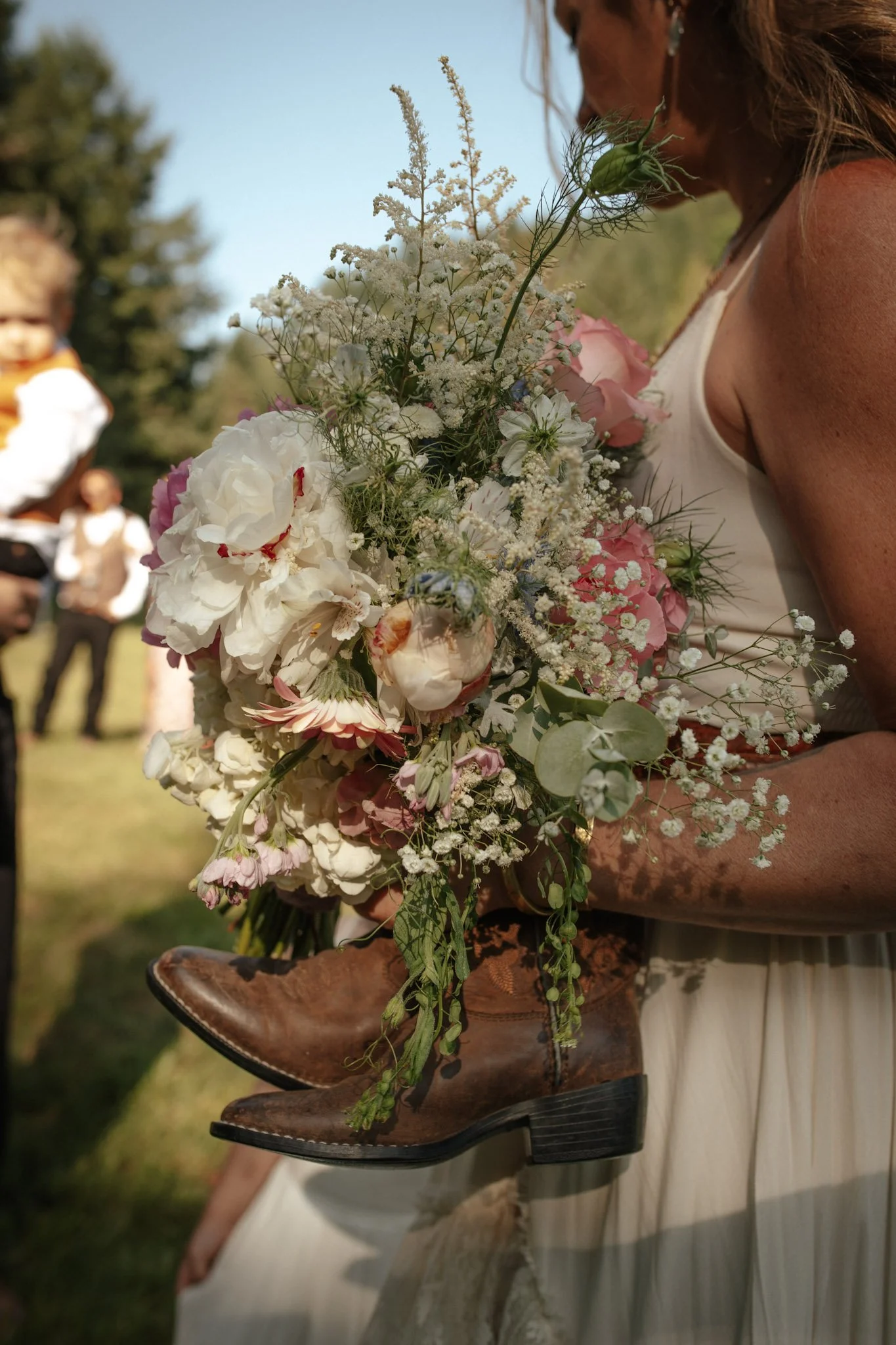 A woman holding a large bouquet of mixed flowers, with a pair of brown cowboy boots hanging from the bouquet. In the background, there are a few people standing outdoors on a sunny day.