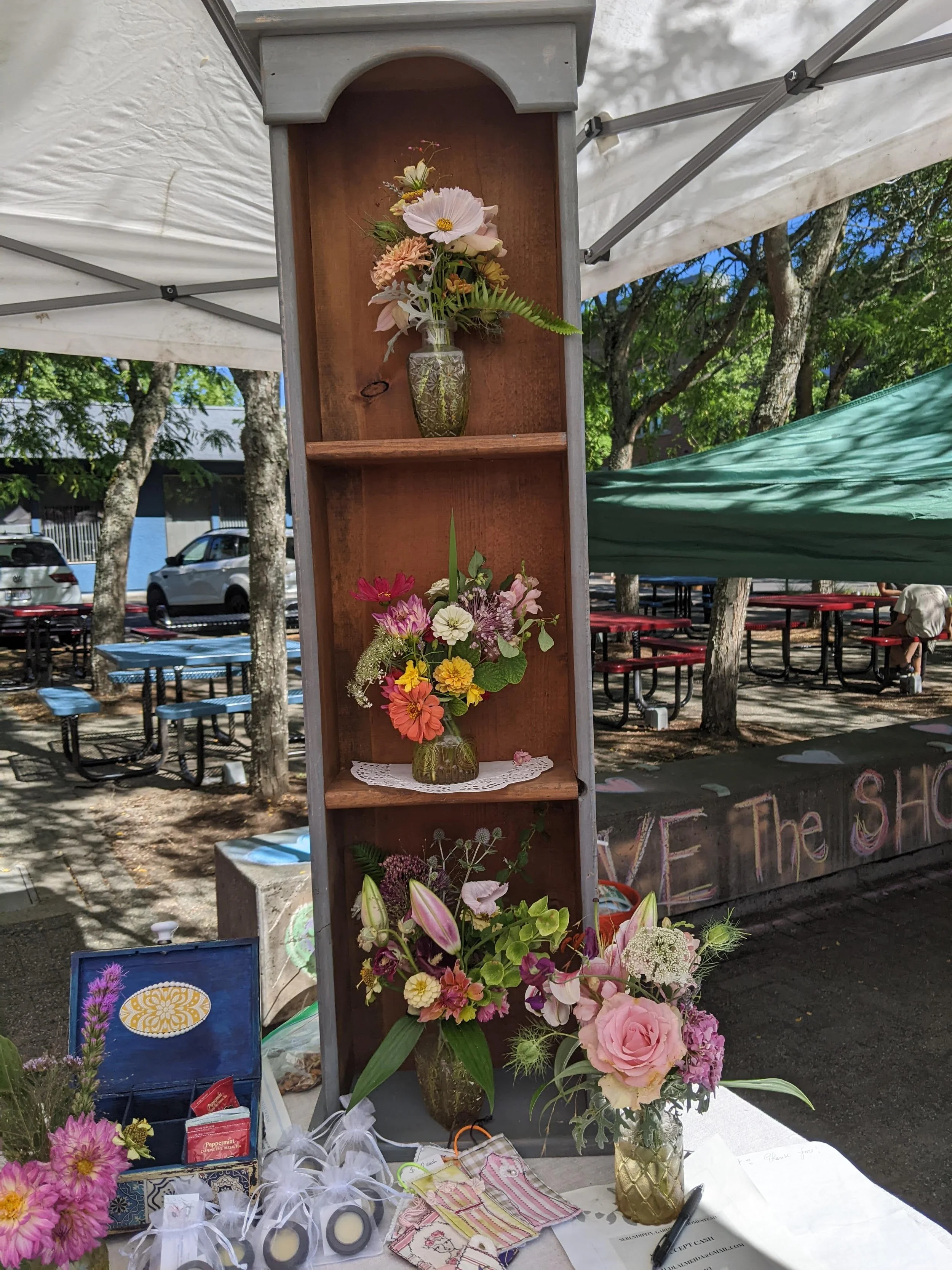 Display of three flower arrangements in vases, set up at an outdoor market stall with additional items including small packages, a pen, and a clipboard on a table. There are trees and picnic tables in the background.