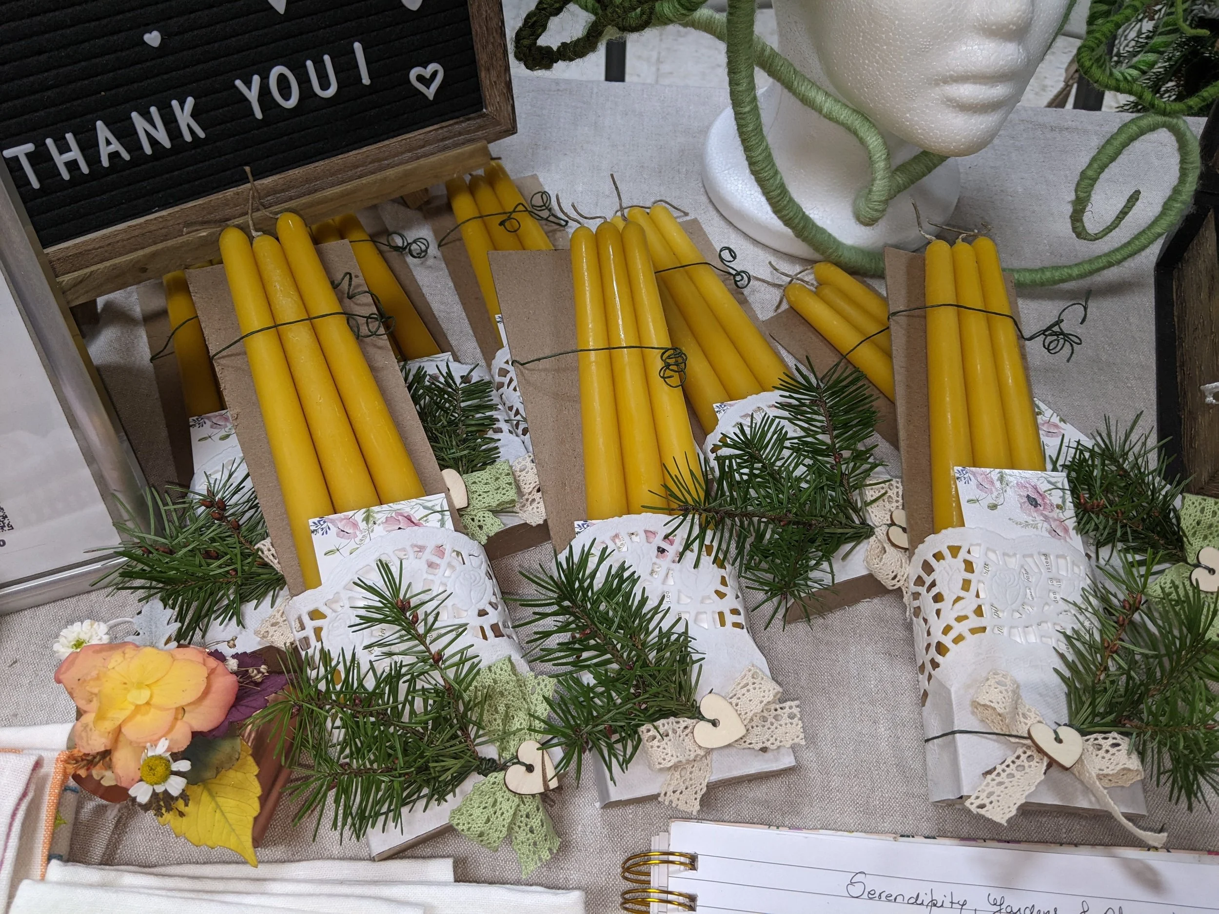 Yellow candles decorated with small pine branches, lace, and paper doilies, arranged on a table with a thankful message board and a decorative head sculpture in the background.