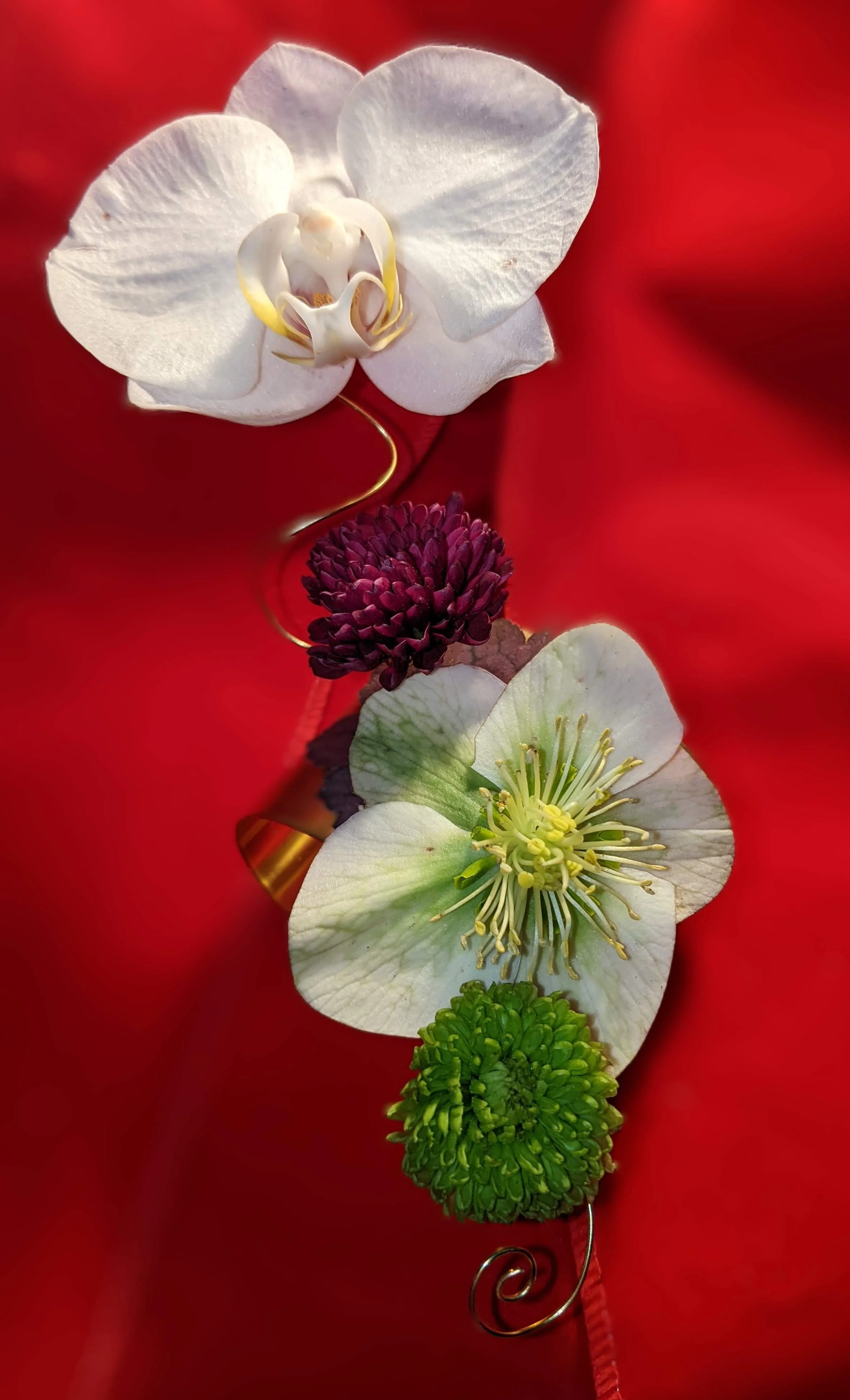 A floral jewelry piece featuring white orchids, a purple globe amaranth, and green pom-pom flower, set against a red fabric background.