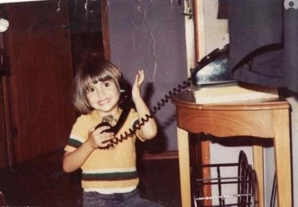 A young girl with a bob haircut smiling while talking on an old corded telephone in a room.