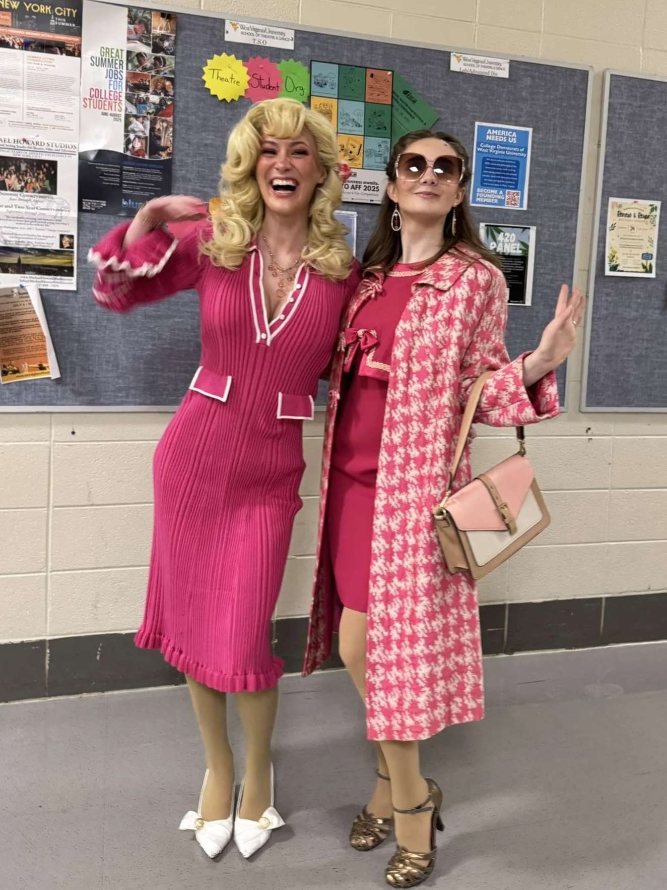 West Virginia University 9 to 5 the Musical. Doralee Rhodes. Two women dressed in vintage pink outfits posing with smiles in front of a bulletin board with various posters and notices.