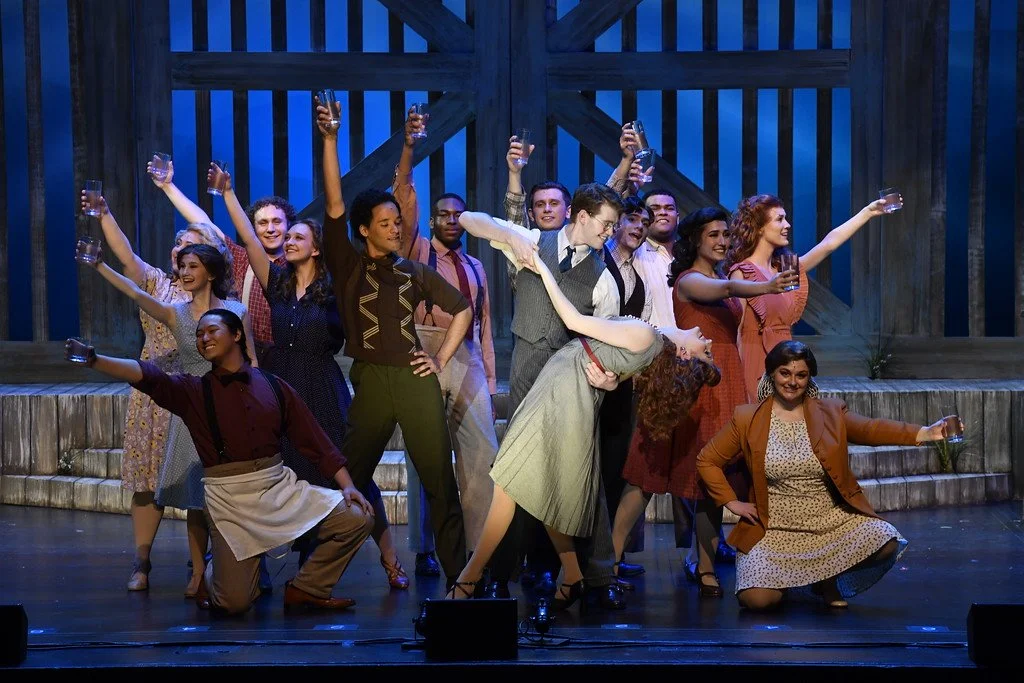 The cast of a theatrical play or musical on stage, celebrating with glasses of drink, in front of a wooden barn backdrop.