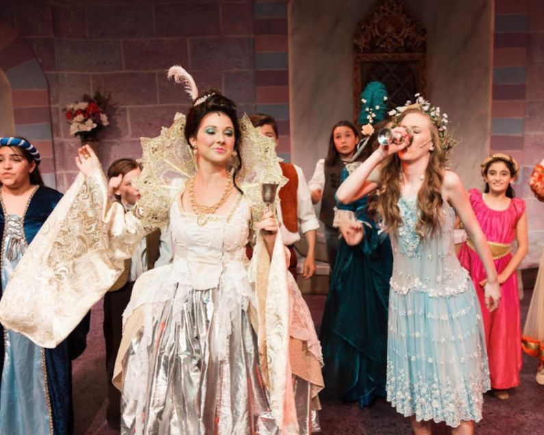Women and children dressed in elaborate costumes, with one woman holding a goblet and another drinking from a glass, in a theatrical setting with a stone wall and decorative background.
