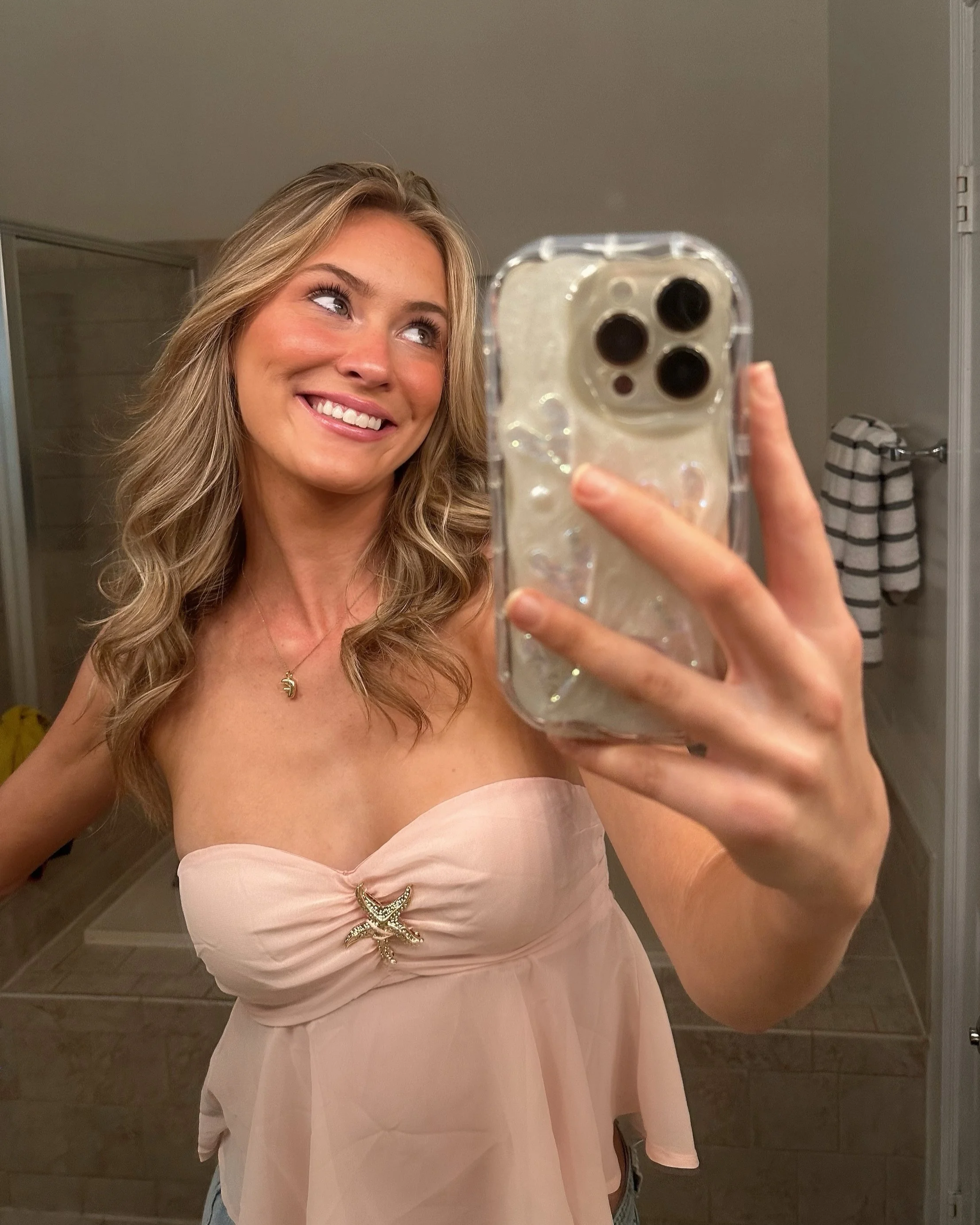 A woman with wavy blonde hair taking a mirror selfie in a bathroom, smiling and wearing a strapless pink dress with a starfish brooch.