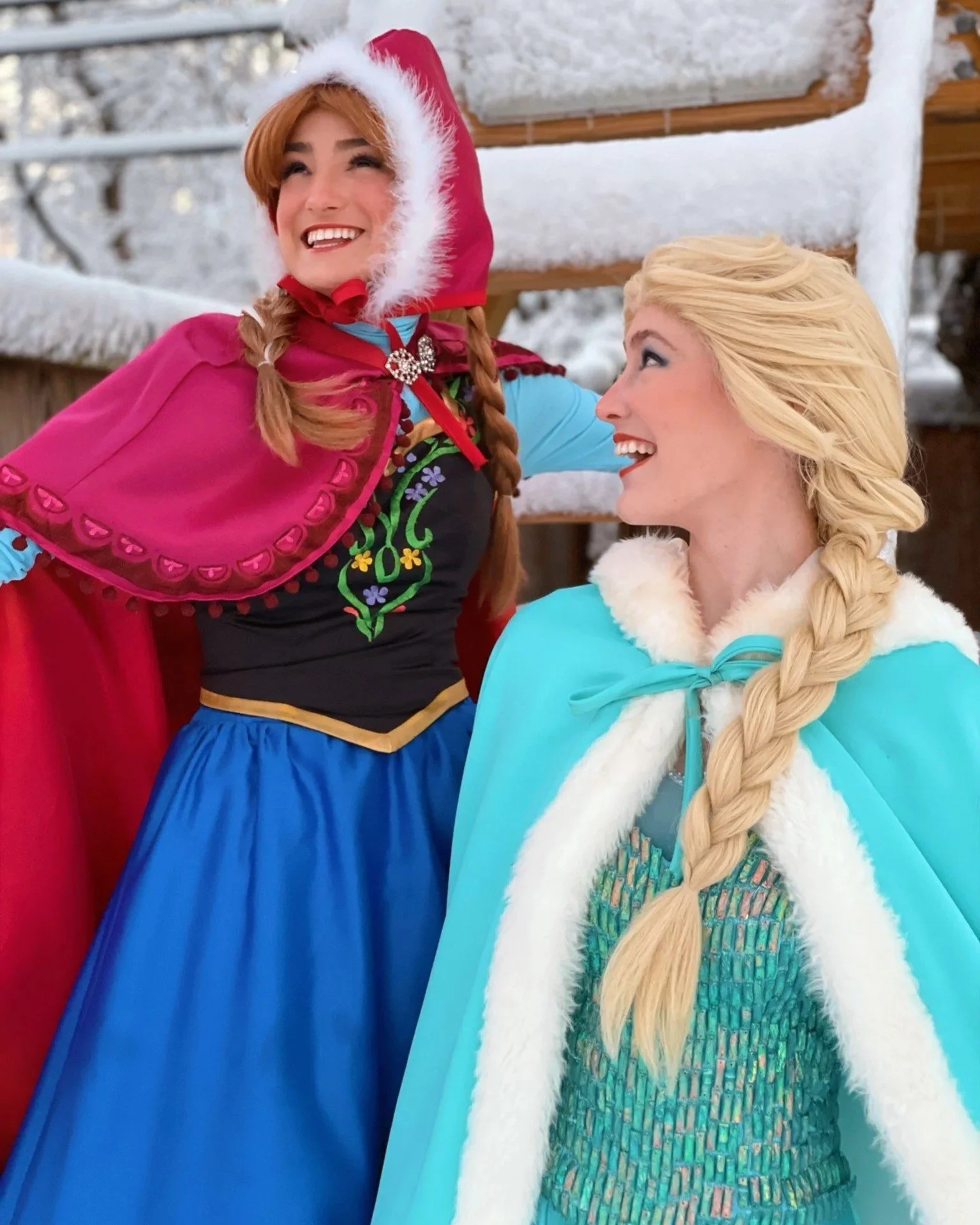 Two women dressed as fairy tale princesses, one in a pink cape and hood, and the other in a light blue cape, standing outdoors in a snowy setting, smiling and looking at each other.