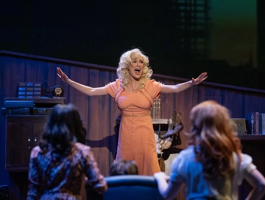 Faith Hargadon as Doralee Rhodes originated by Dolly Parton in 9 to 5 the Musical at West Virginia University. A woman with curly blonde hair wearing an orange dress is on stage, gesturing with her arms outstretched, 
