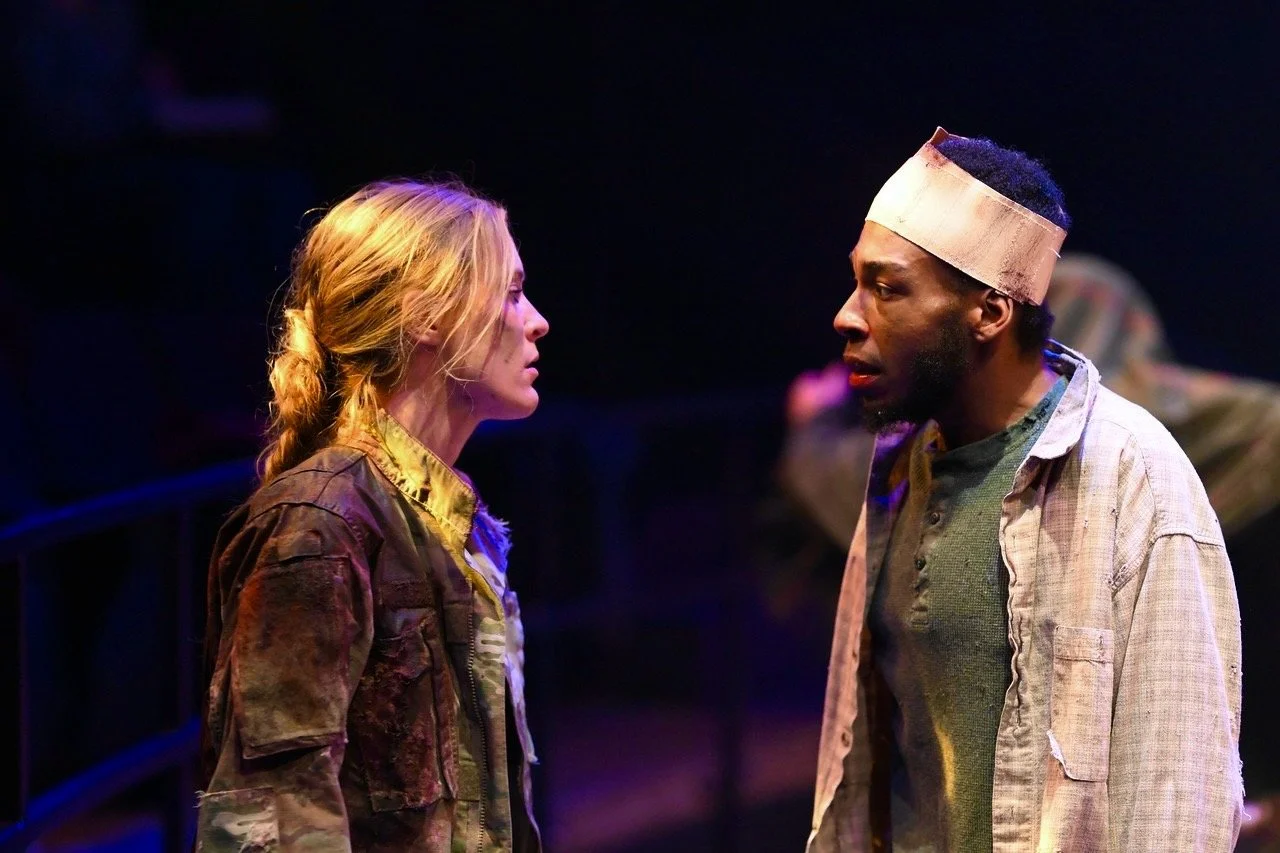 Two actors on stage facing each other in a theatrical performance, one woman with blonde hair in a camouflage jacket and one man with a headband and casual clothing, engaged in a serious conversation.