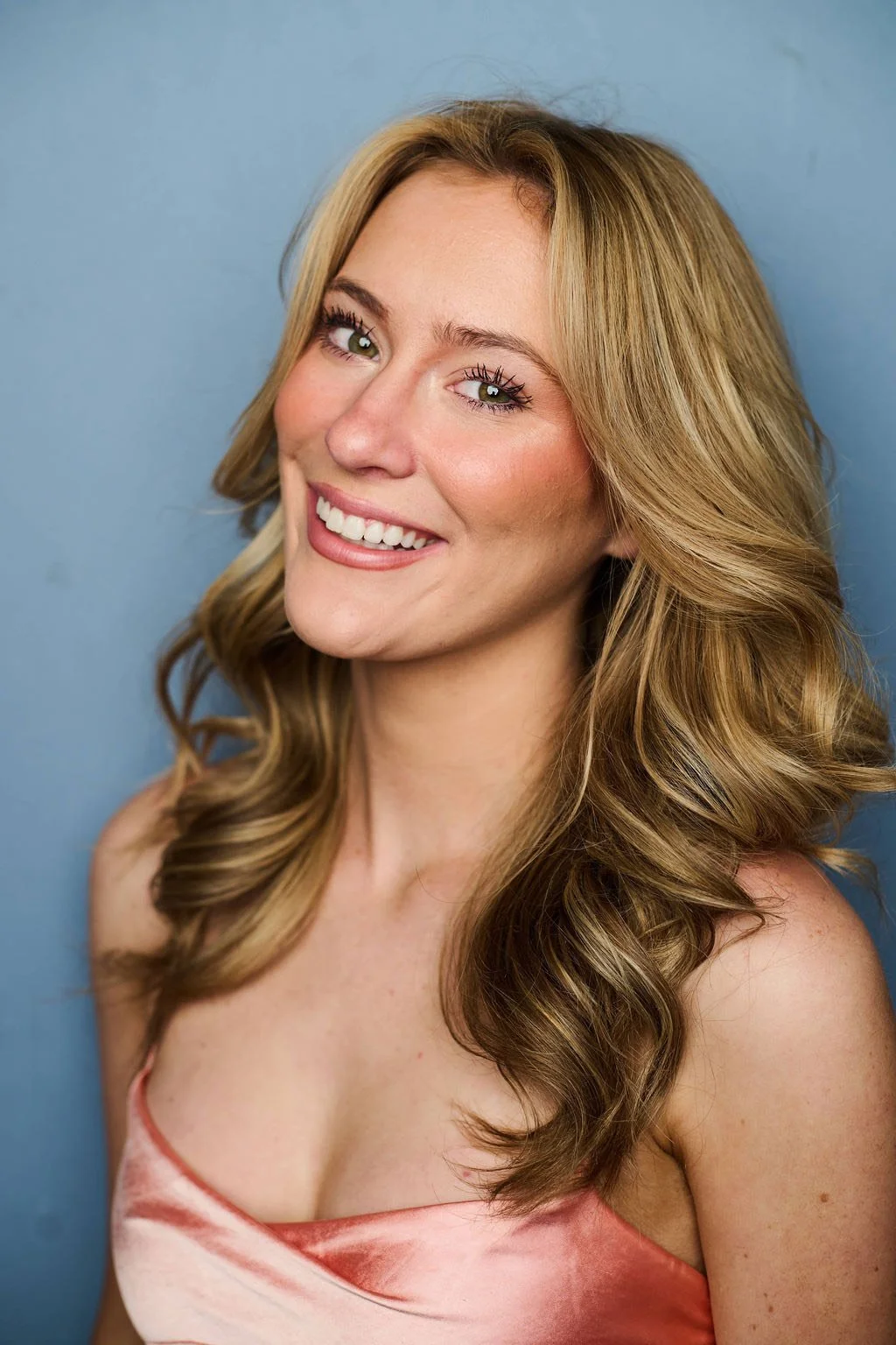 Faith Hargadon theatrical musical theatre headshot. A smiling woman with long wavy blonde hair, light eye makeup, and a pink satin sleeveless top, standing against a light blue background.