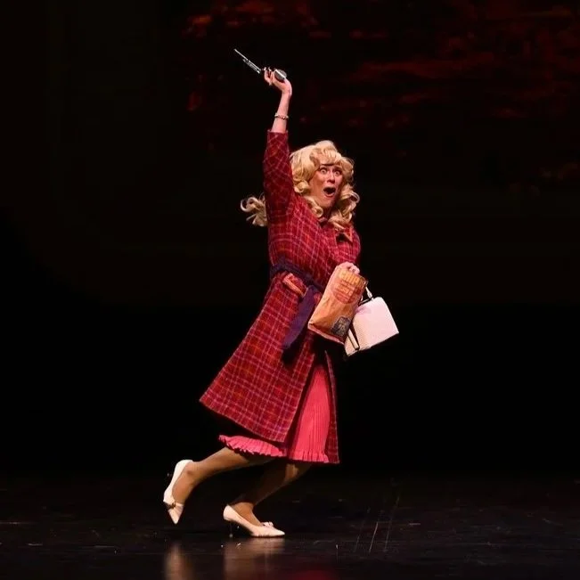 A woman dressed in a vintage pink and red plaid outfit with a coat, skirt, and heels, holding a syringe raised high, with a surprised or intense facial expression, on a dark stage background.