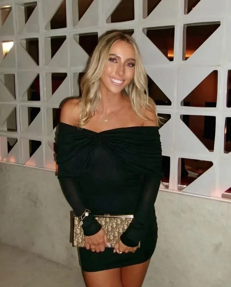 A woman with blonde hair and a big smile wearing a black off-the-shoulder dress and long black gloves, holding a patterned clutch purse, standing indoors against a white geometric patterned wall.
