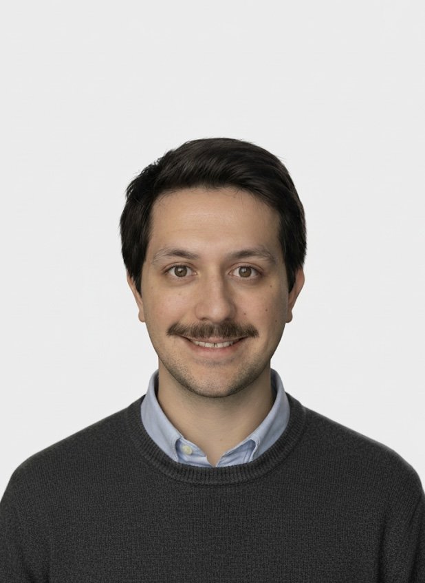 A young man with dark hair, a mustache, and light skin, wearing a light blue collared shirt and dark sweater, smiling against a plain white background.