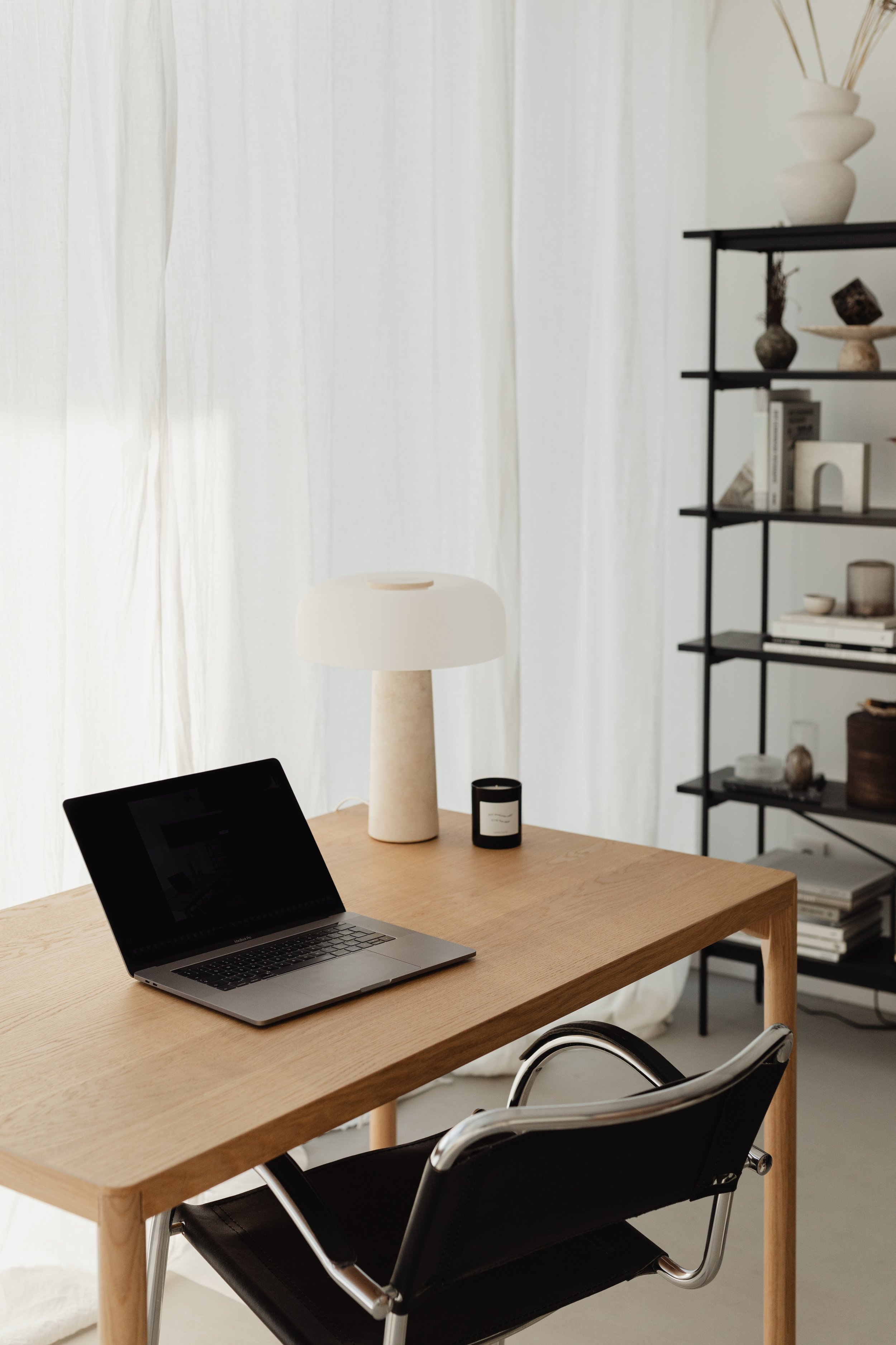 A minimalist home office with a wooden desk, a laptop, a modern white table lamp, and a black metal bookshelf filled with decorative items and books, set in front of white curtains.