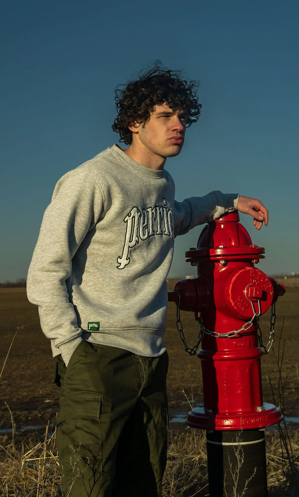 A young man with curly hair, wearing a gray sweatshirt and green cargo pants, standing outdoors and leaning on a red fire hydrant in a vast field under a clear blue sky.
