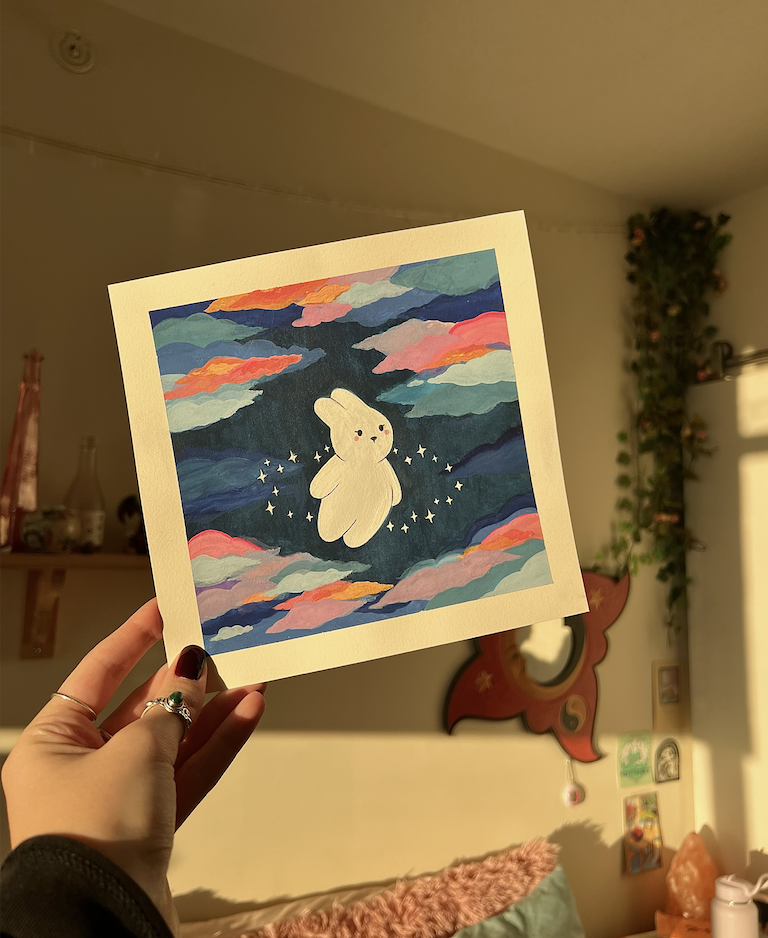 Hand holding a colorful illustration of a white bunny surrounded by stars, floating among clouds at sunset.