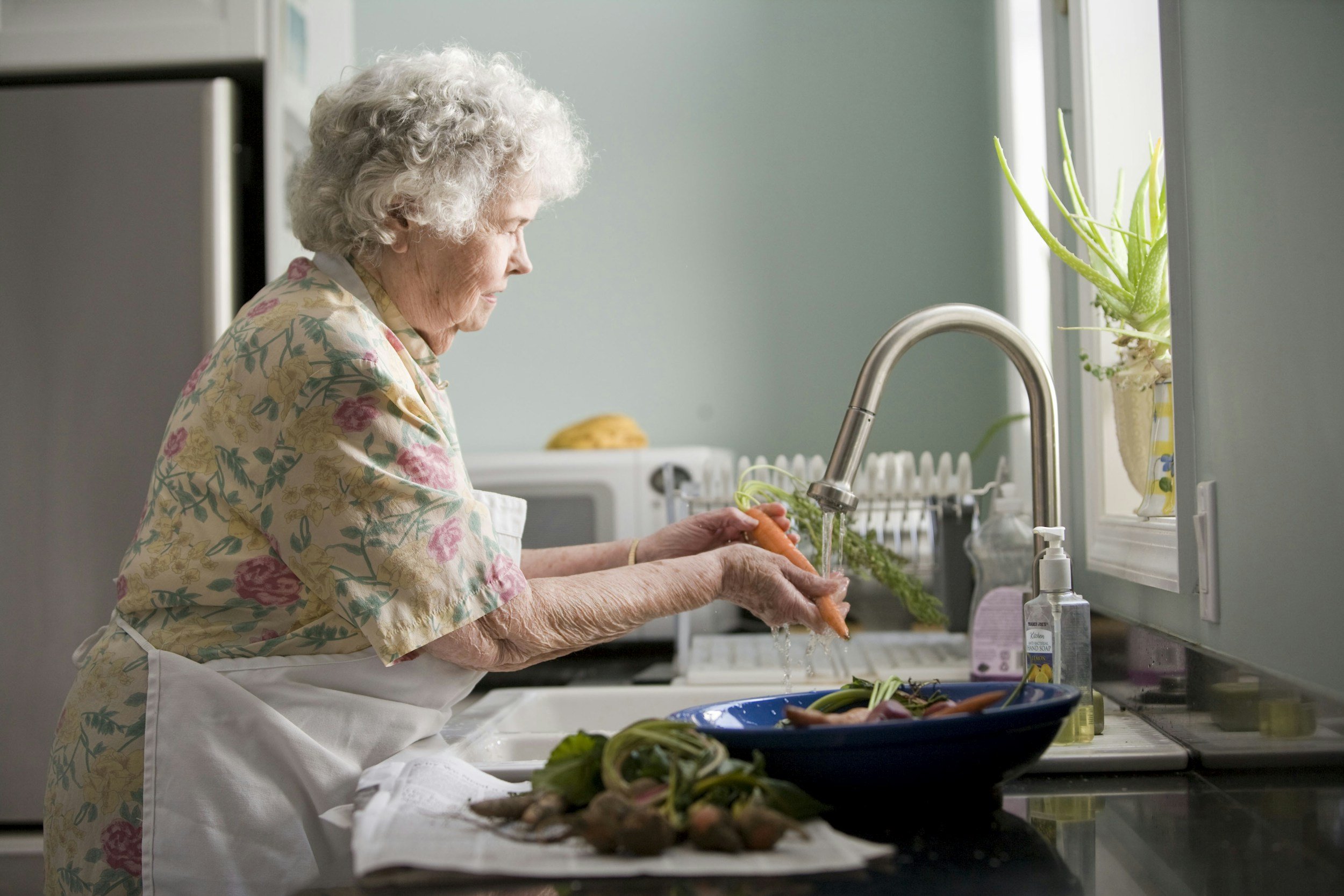 An elderly woman with curly gray hair washing carrots at the kitchen sink.