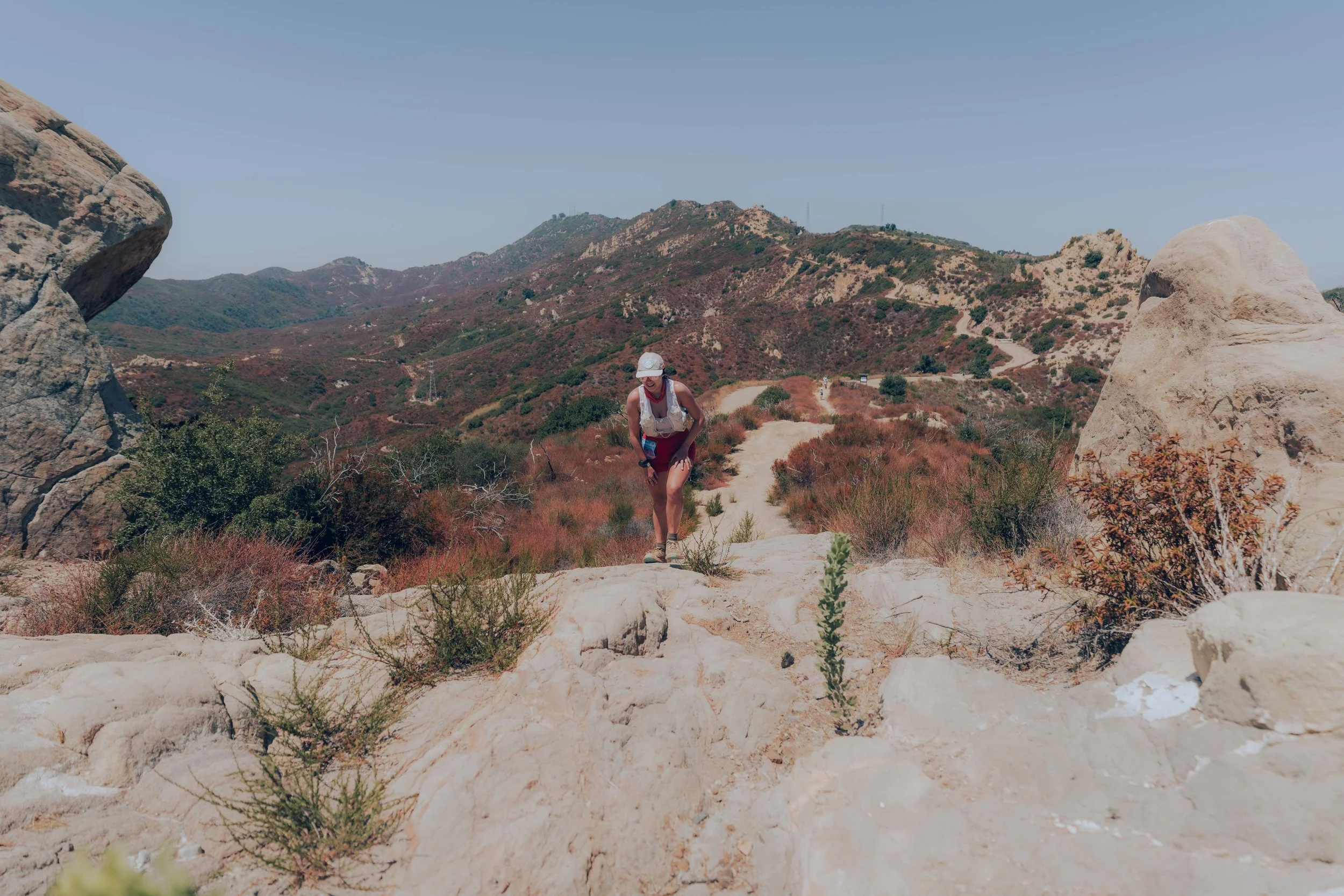 Hiker wearing a white hat, sunglasses, and red shorts walks on a rocky trail in a mountainous area with sparse vegetation and large boulders.