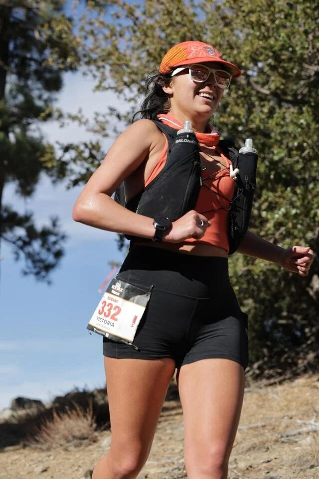 Female runner in athletic gear, including sunglasses, a cap, and a hydration vest, participating in a trail race outdoors.