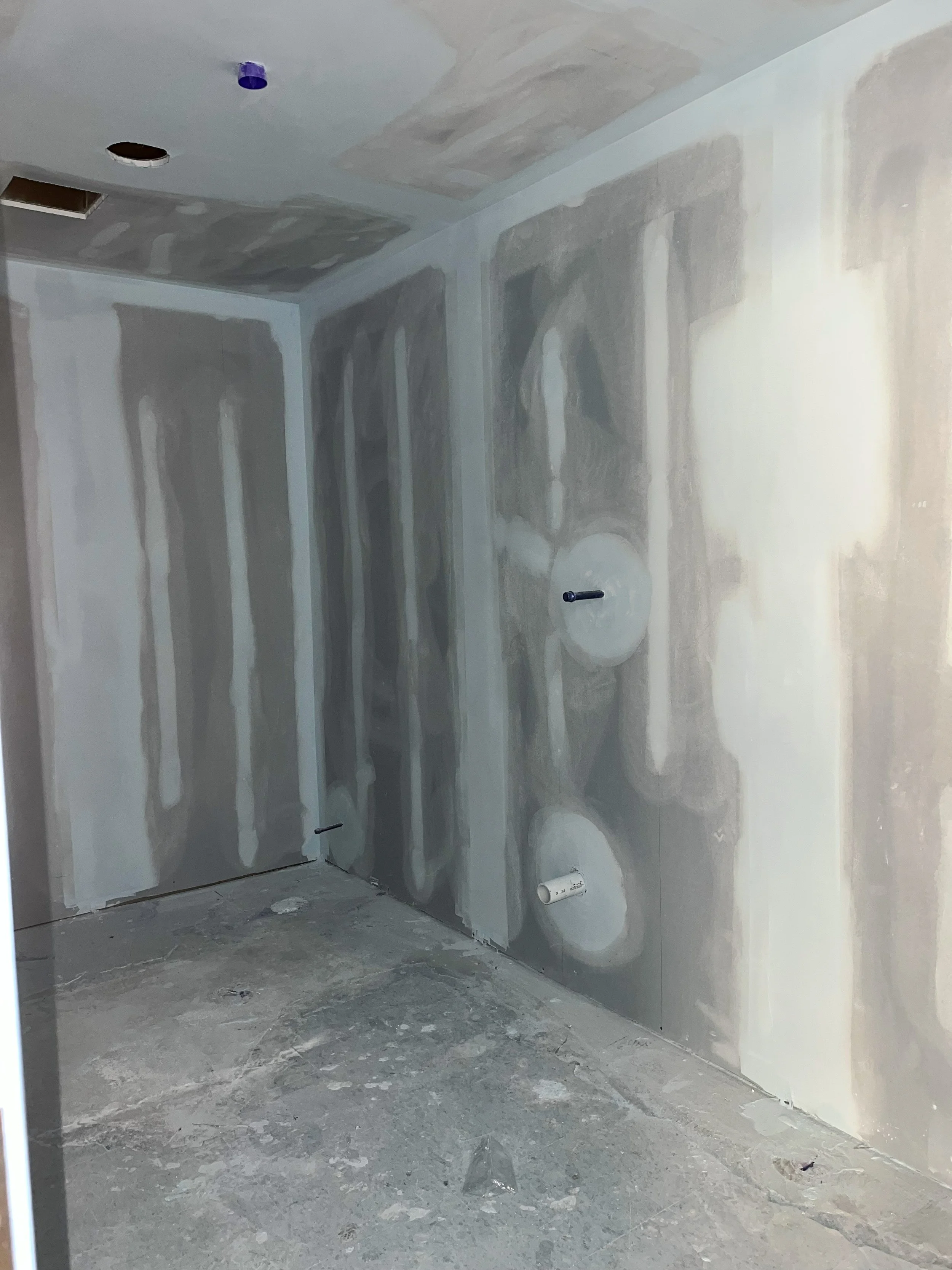 Drywall installation in a bathroom with plumbing pipes protruding from the wall, and an exhaust vent opening in the ceiling.