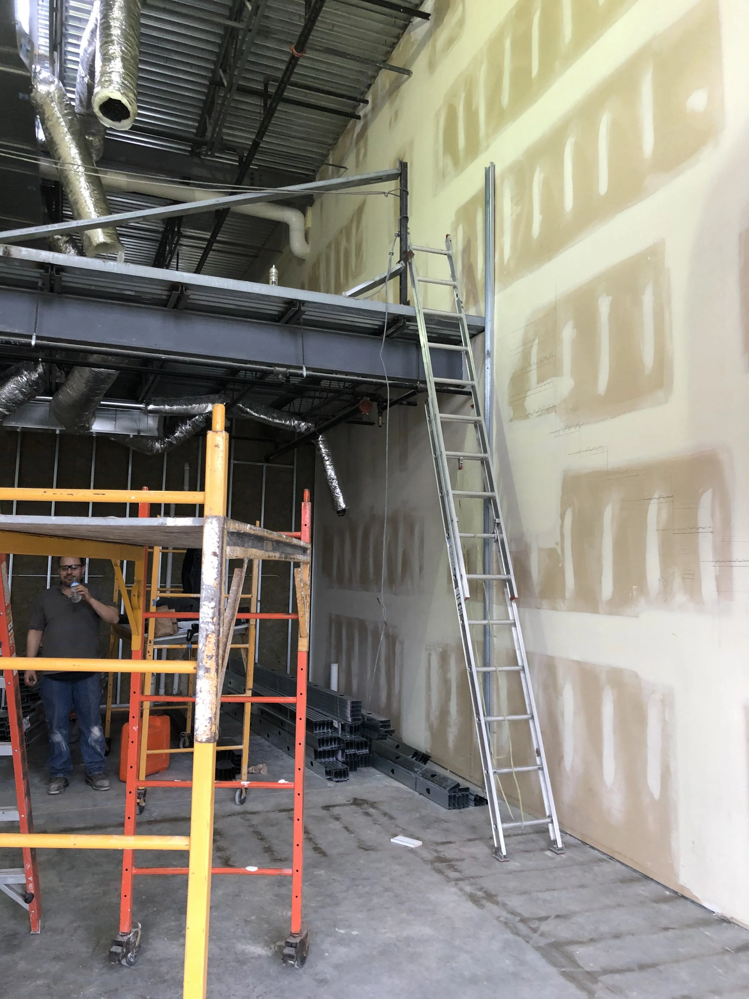 Interior of a construction site with scaffolding, a ladder, and partially finished wall drywall. A man is standing near the scaffolding, drinking water.