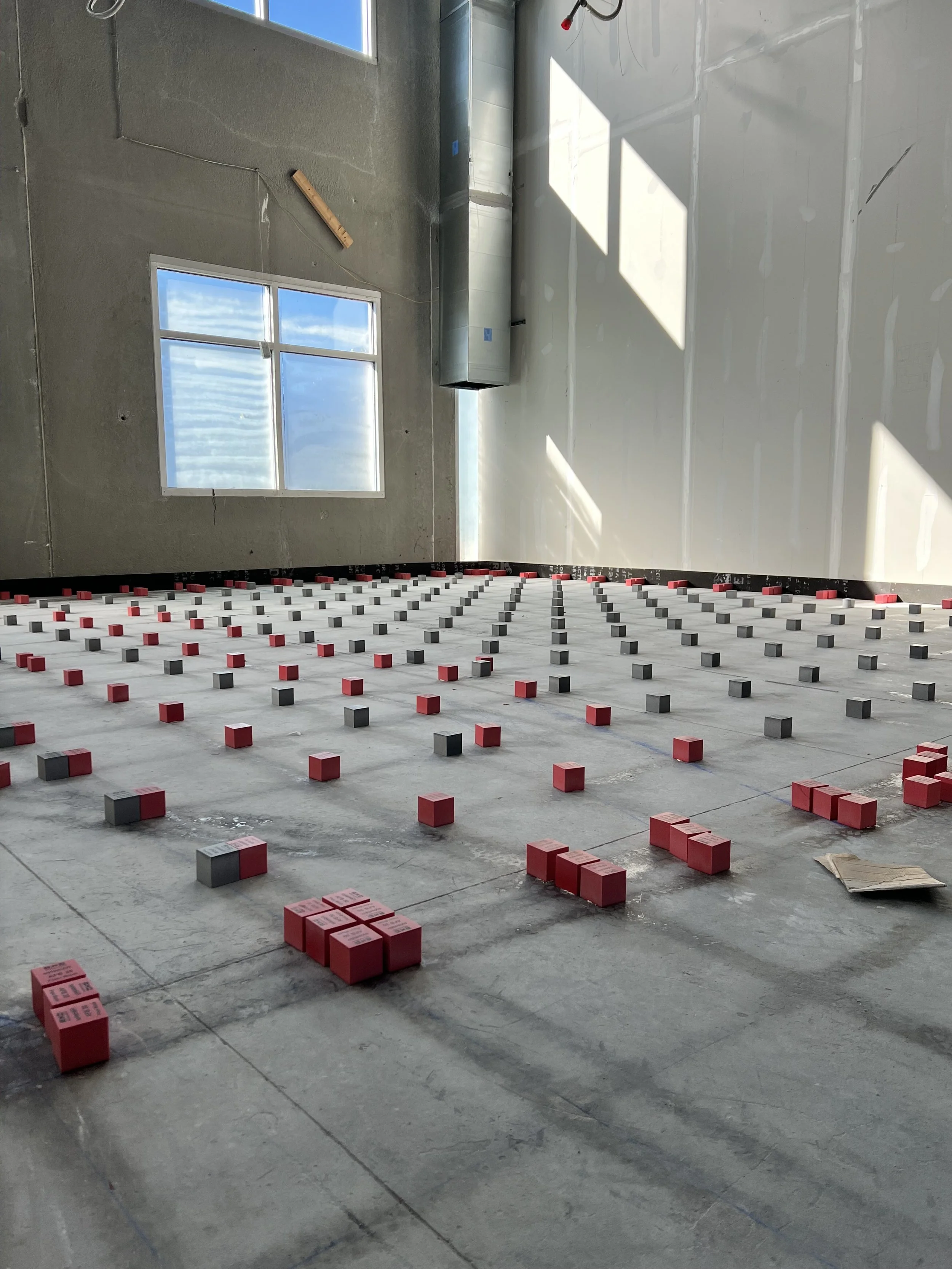 Interior of a construction site with a concrete floor underlay, with red and black tile spacers arranged in a grid for a flooring project. There is a large window on the wall providing natural light, and construction tools and ductwork are visible.