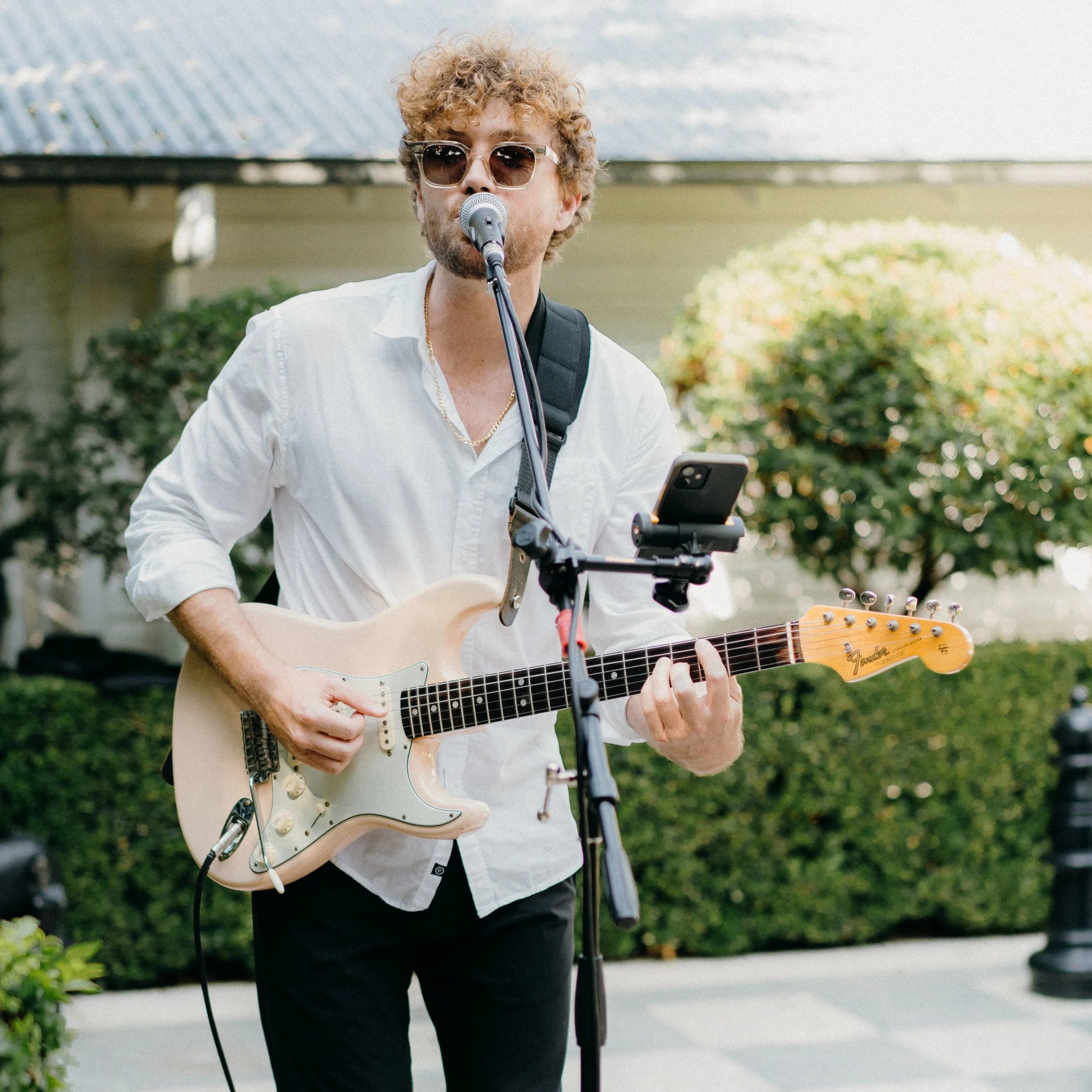 A young man with curly blonde hair, wearing sunglasses and a white shirt, is playing an electric guitar while singing into a microphone outdoors.