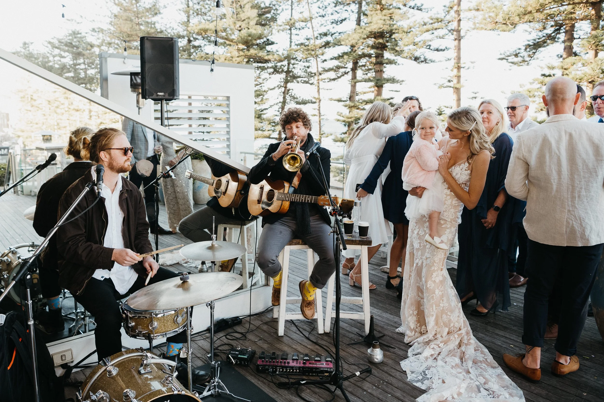 Musicians playing instruments at a wedding reception while guests dance and socialize outdoors surrounded by trees.