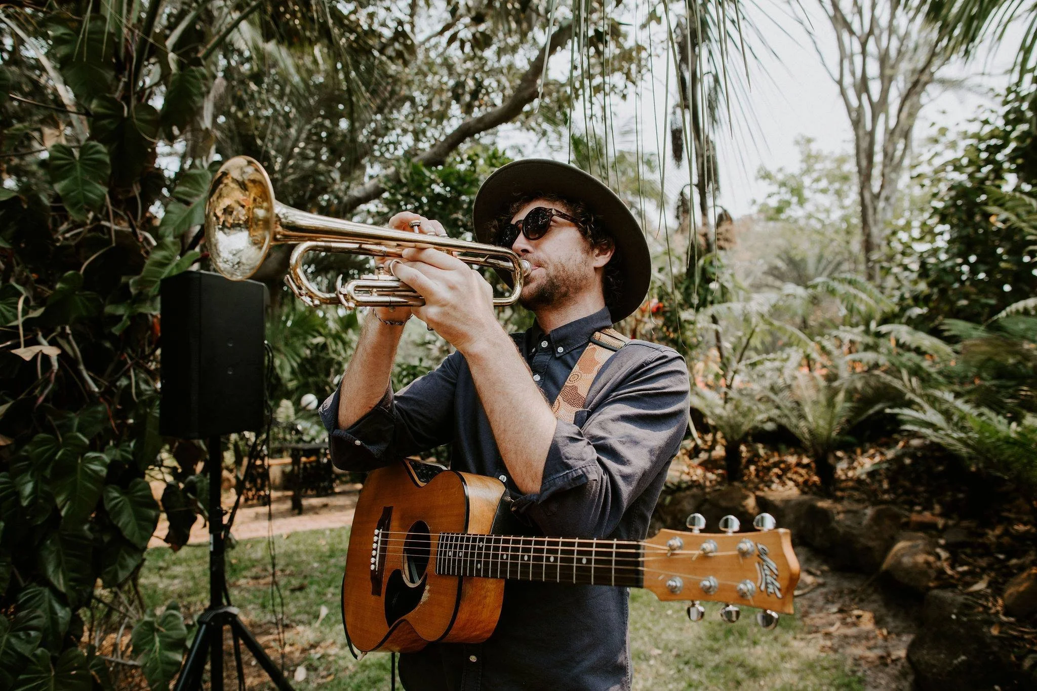 A man wearing sunglasses and a hat plays a trumpet and holds an acoustic guitar with a strap around his shoulder outdoors in a lush, green garden.