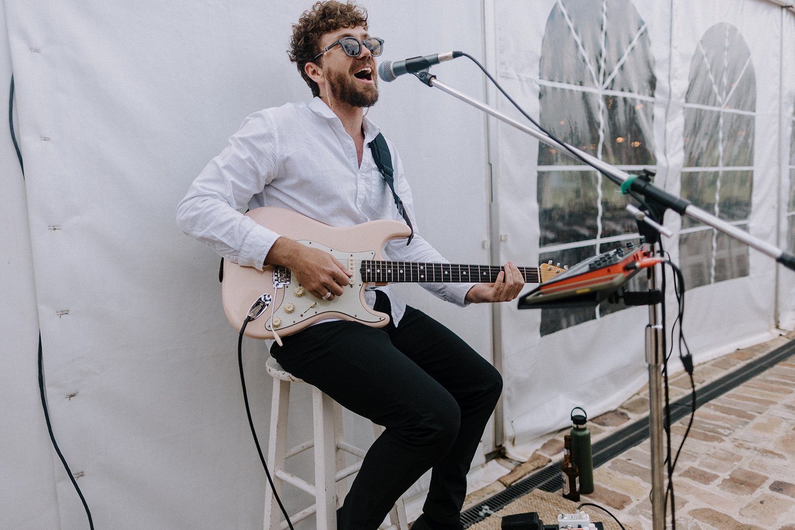 A man with curly hair, glasses, and a beard, wearing a white shirt, singing into a microphone while playing an electric guitar. He is seated on a stool, behind a music stand with electronic equipment, in an indoor setting with white walls and large w