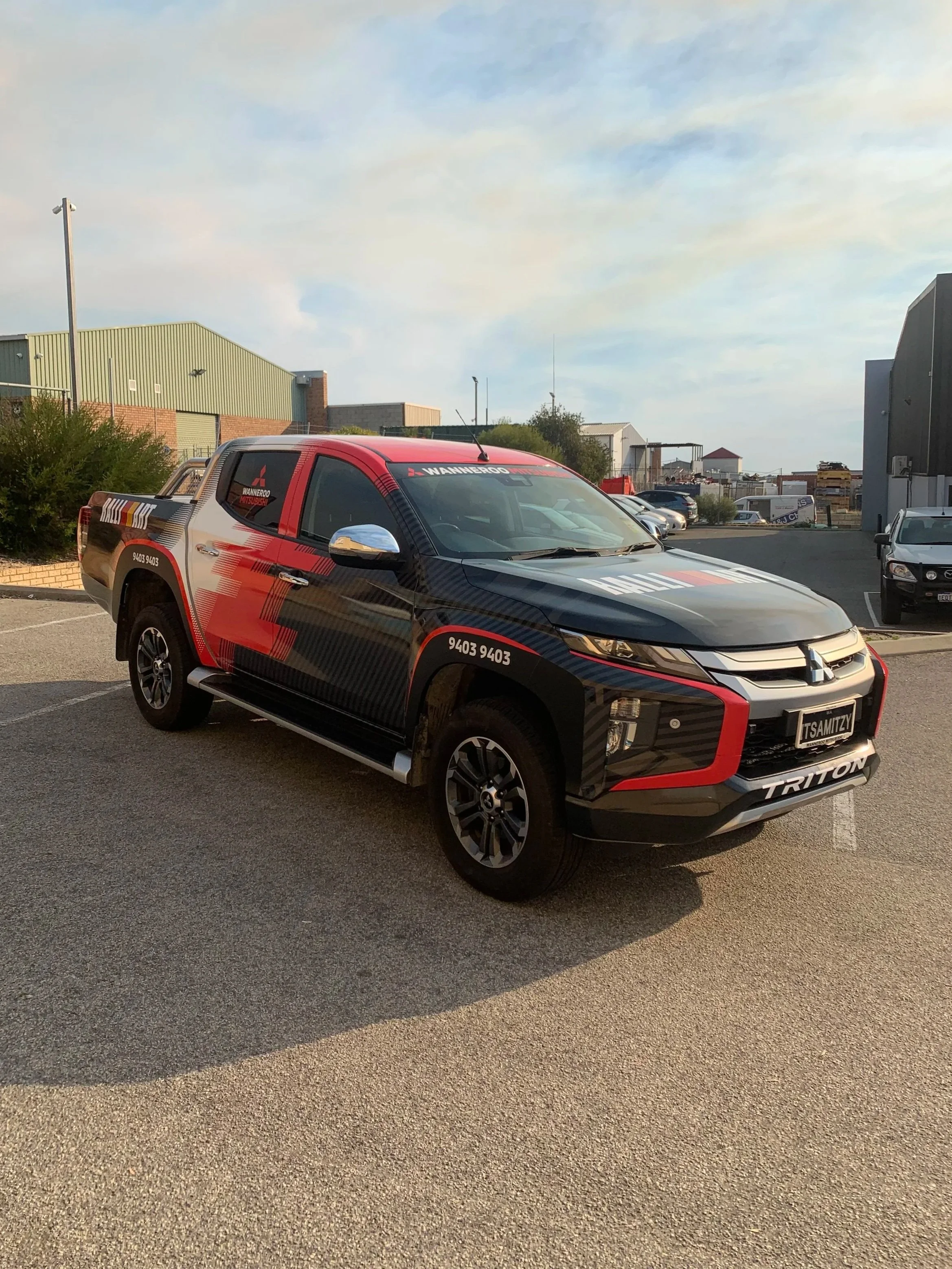 A black and red Mitsubishi Triton pickup truck parked in an outdoor parking lot, with buildings and other vehicles in the background under a partly cloudy sky.