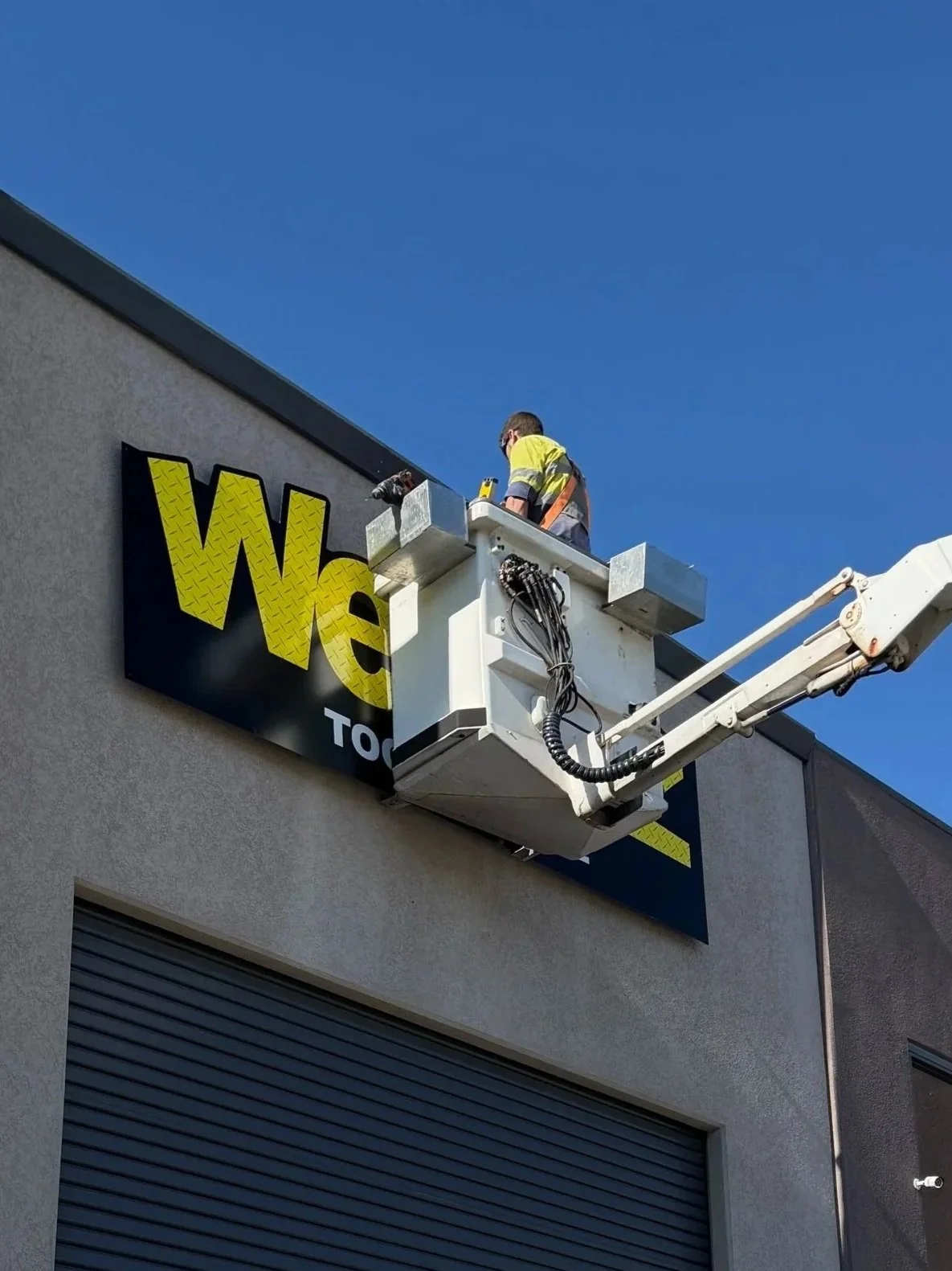 Worker installing or repairing a sign on a building using a cherry picker. The sign reads 'We' and part of another word is visible.