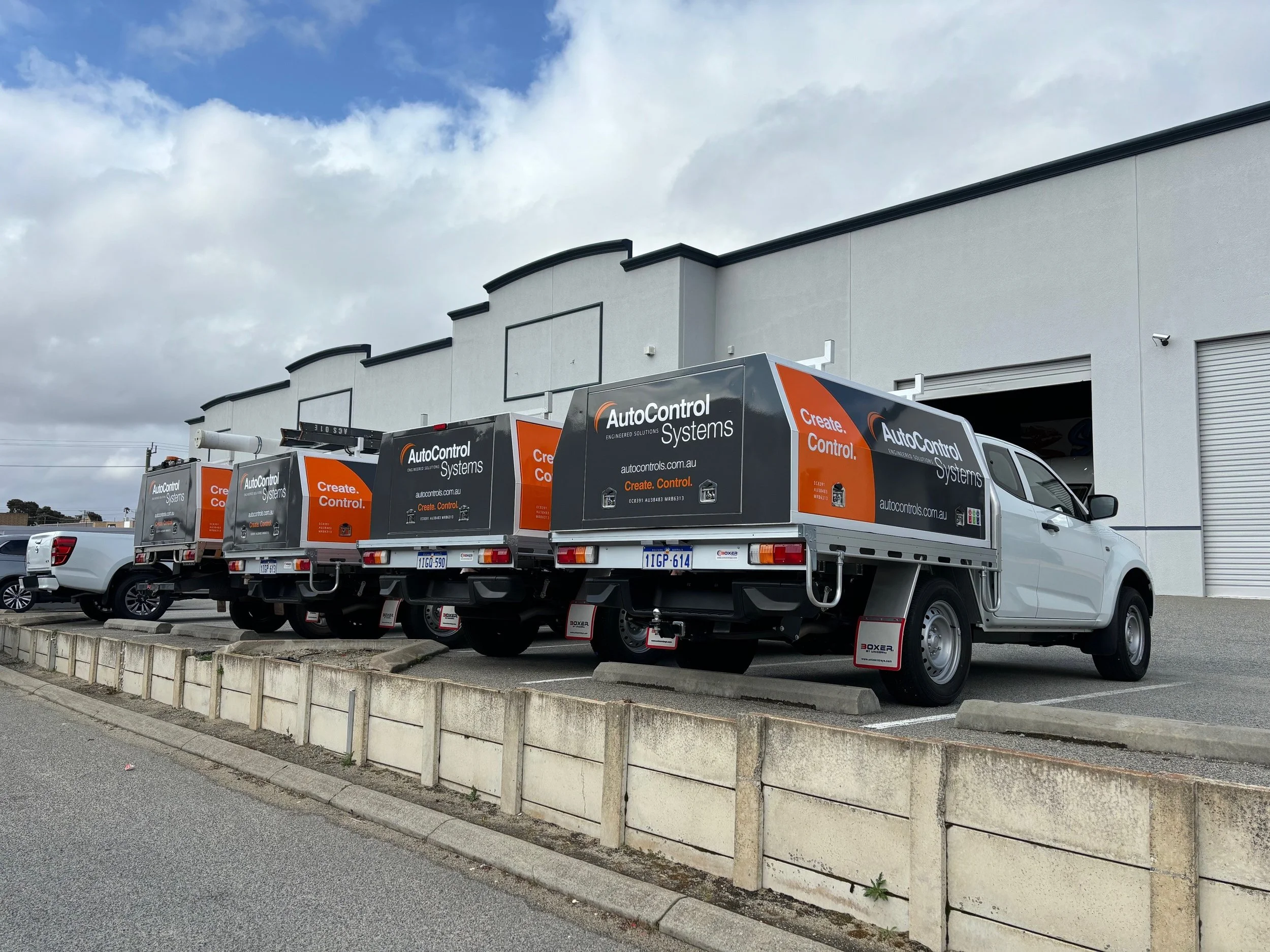 Four trucks with orange and black advertising for AutoControl Systems, parked side by side in front of a white building with closed garage doors.