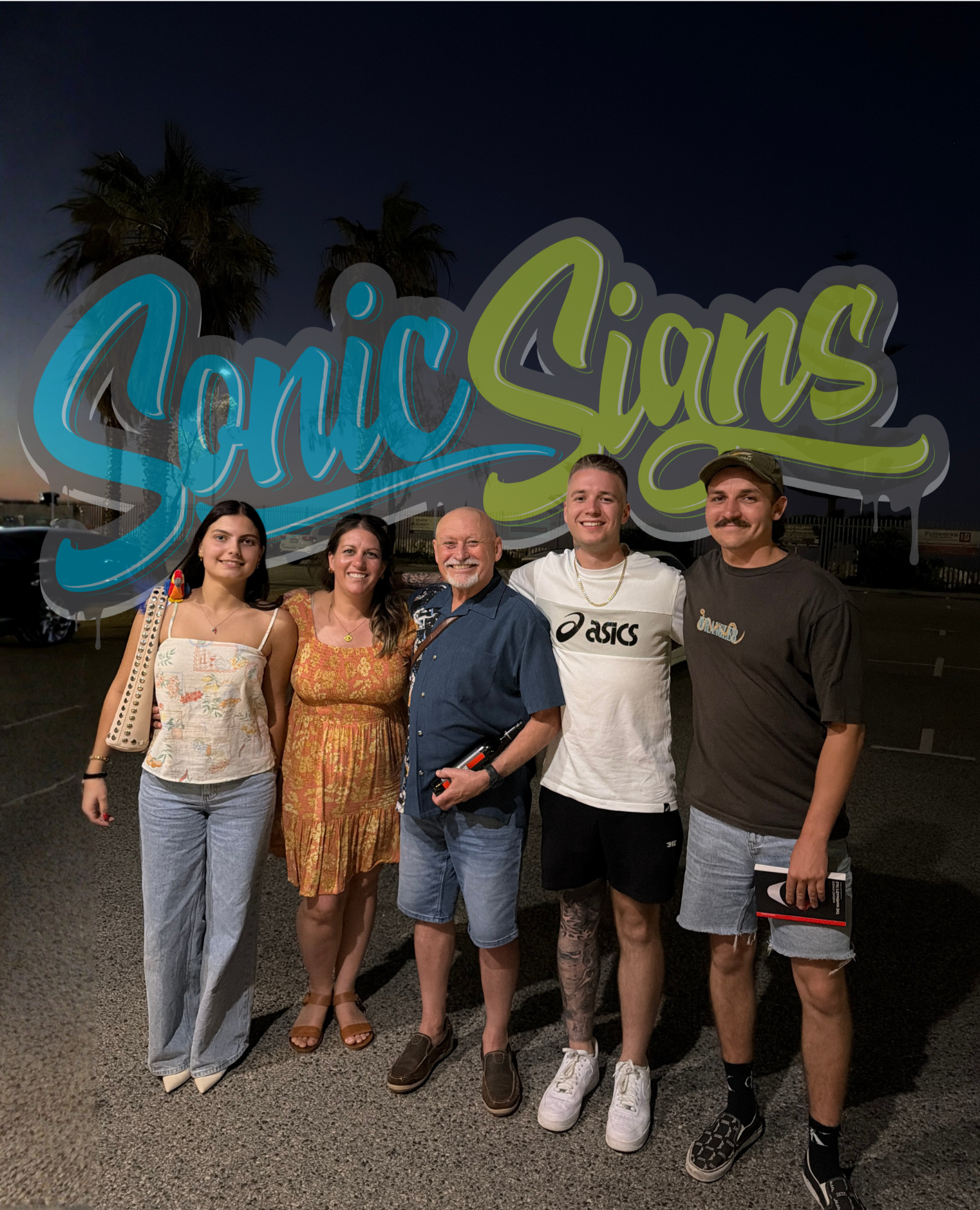 Five people posing in front of a large colorful sign that reads 'Genic Arts' in an outdoor area during the evening. The group includes three men and two women, all smiling, dressed casually, with palm trees visible in the background.