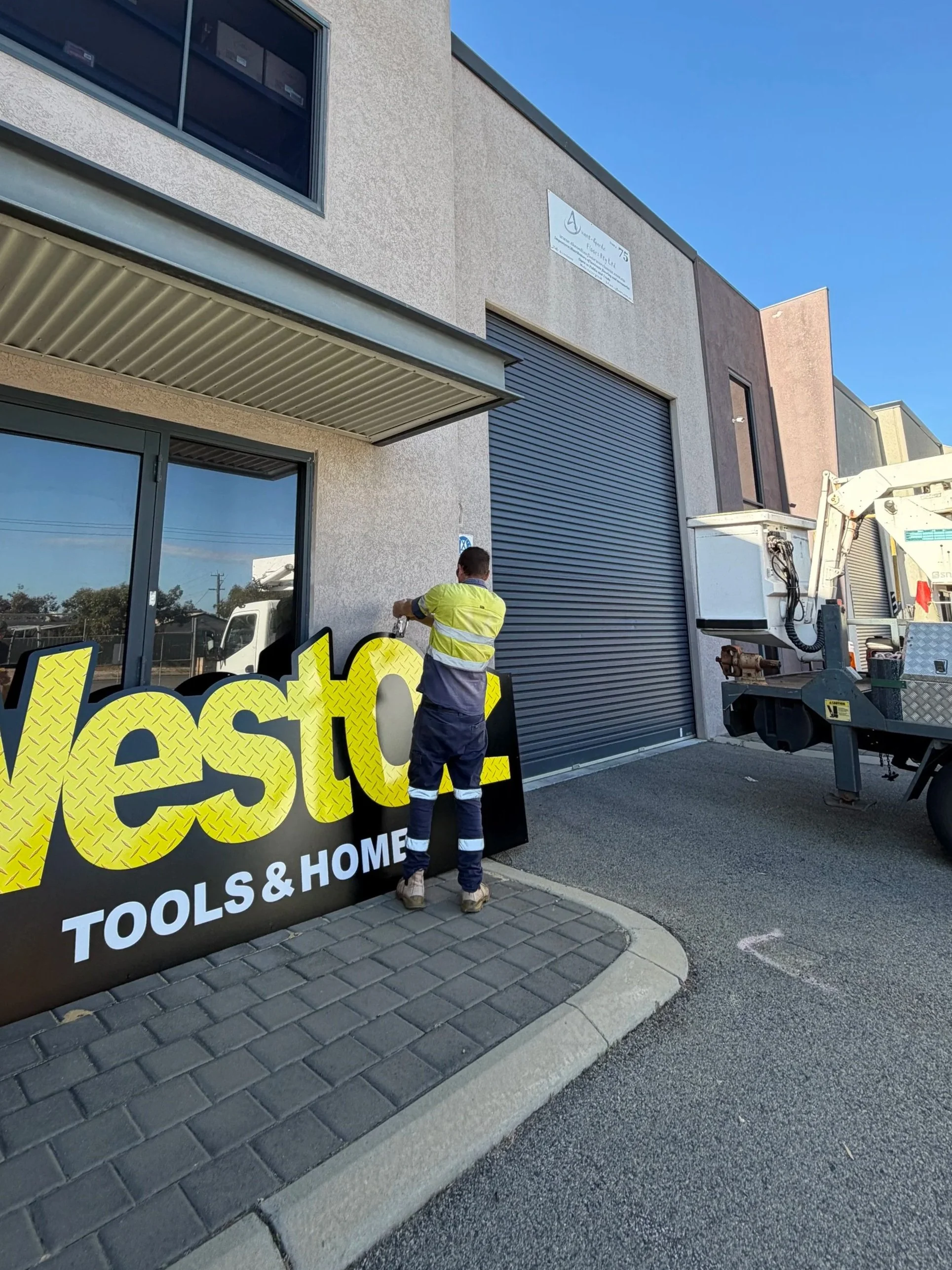 Construction worker setting up a sign outside a building with a gray roller shutter door, large windows, and a sign on the wall; a utility truck and construction equipment are nearby.