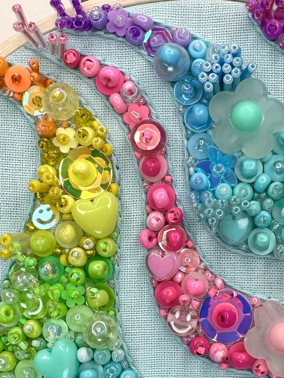 Colorful bead embridery with pink, yellow, blue, purple, and green beads, including heart-shaped and flower-shaped beads, laid out on a textured fabric background.