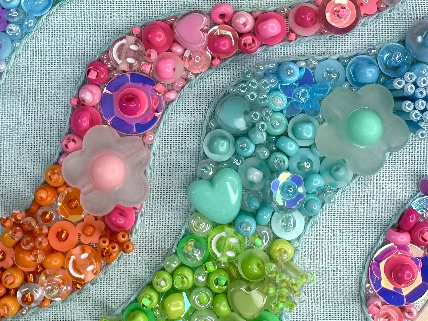 Colorful beaded embroidery with pink, orange, green, blue, and purple beads and floral-shaped beads, displayed on a textured surface.