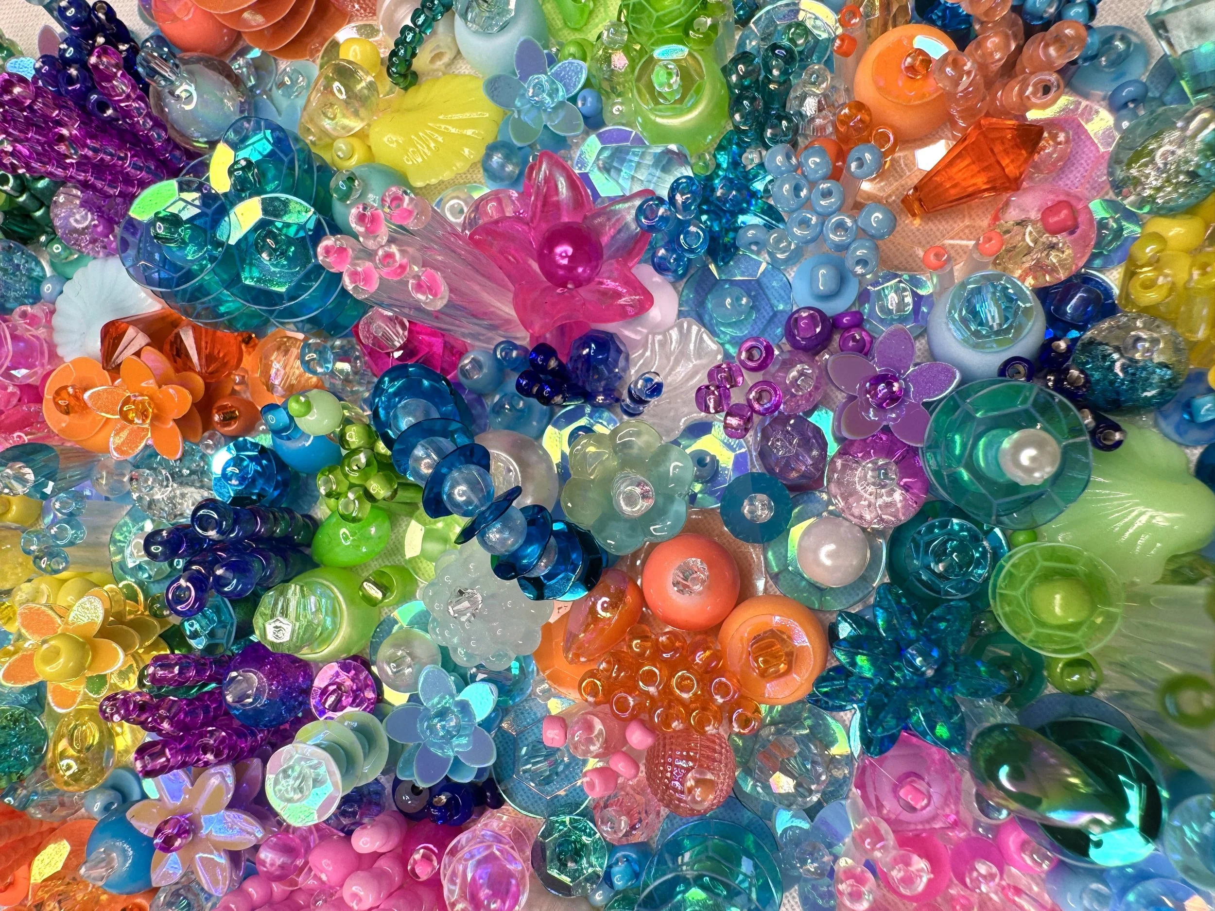 A large collection of colorful plastic beads and charms in various shapes, sizes, and colors including pink, blue, yellow, green, orange, and purple, with some beads resembling flowers and others being geometric or abstract shapes.