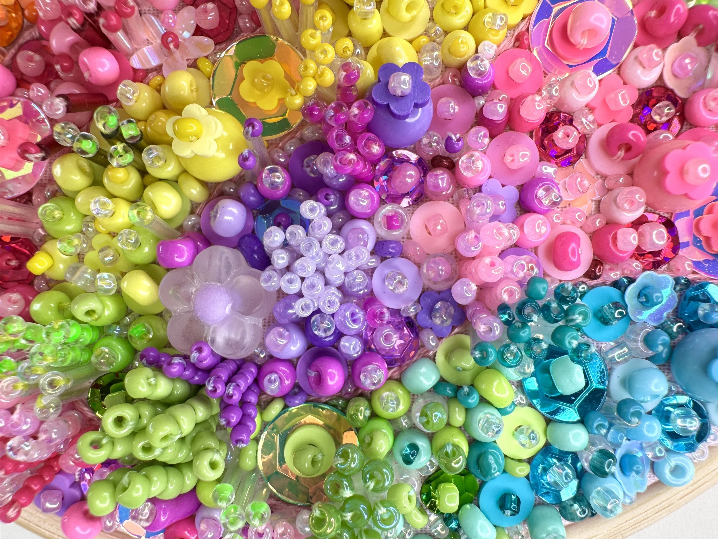 A close-up of colorful plastic beads and embellishments, including pink, yellow, purple, green, and blue beads, some shiny and some matte, arranged in a pattern. Some beads are shaped like flowers, and there are reflective and holographic pieces scat