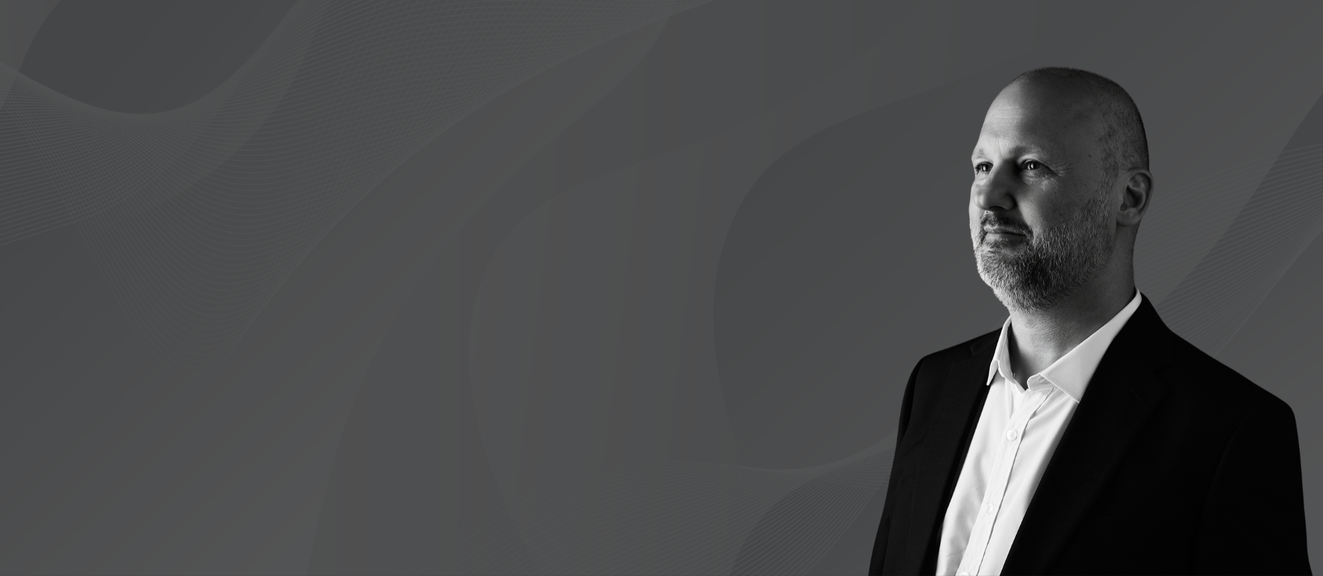 Black and white portrait of a man in a suit with a beard, looking to the right, against a dark, abstract geometric background.