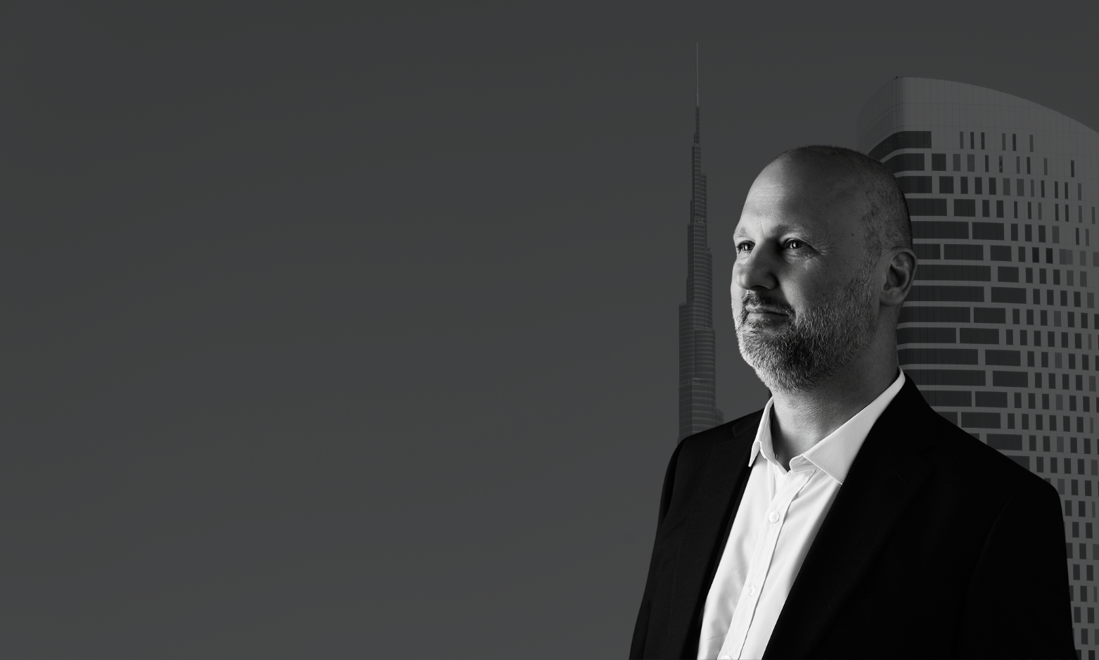 A black and white photo of a middle-aged man with a beard, dressed in a suit, looking to the right, with a city skyline featuring skyscrapers in the background.