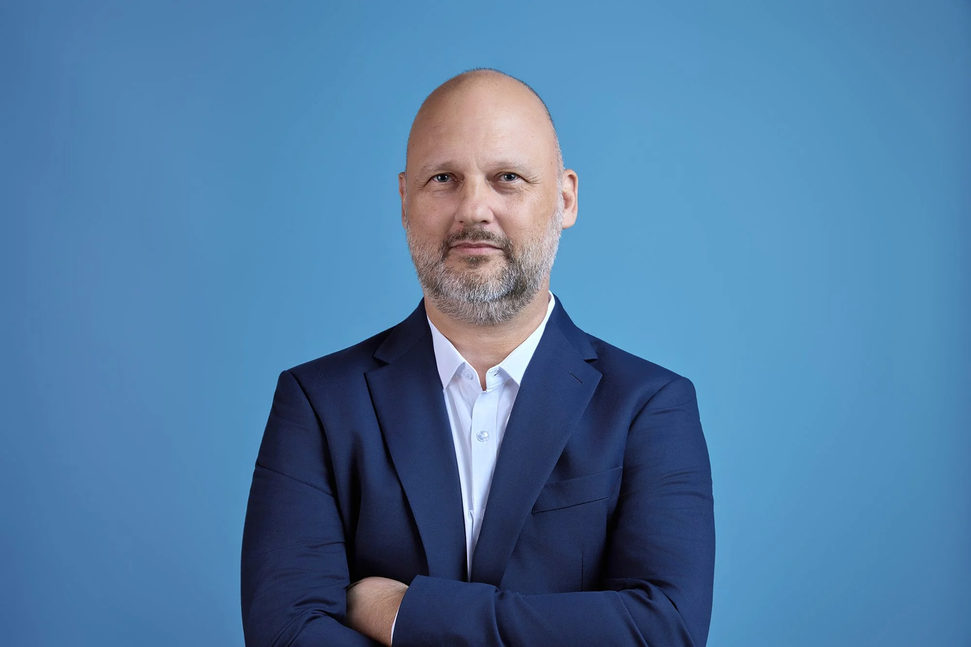 A middle-aged man with a bald head and gray beard, wearing a navy blazer and white shirt, standing against a light blue background with his arms crossed.