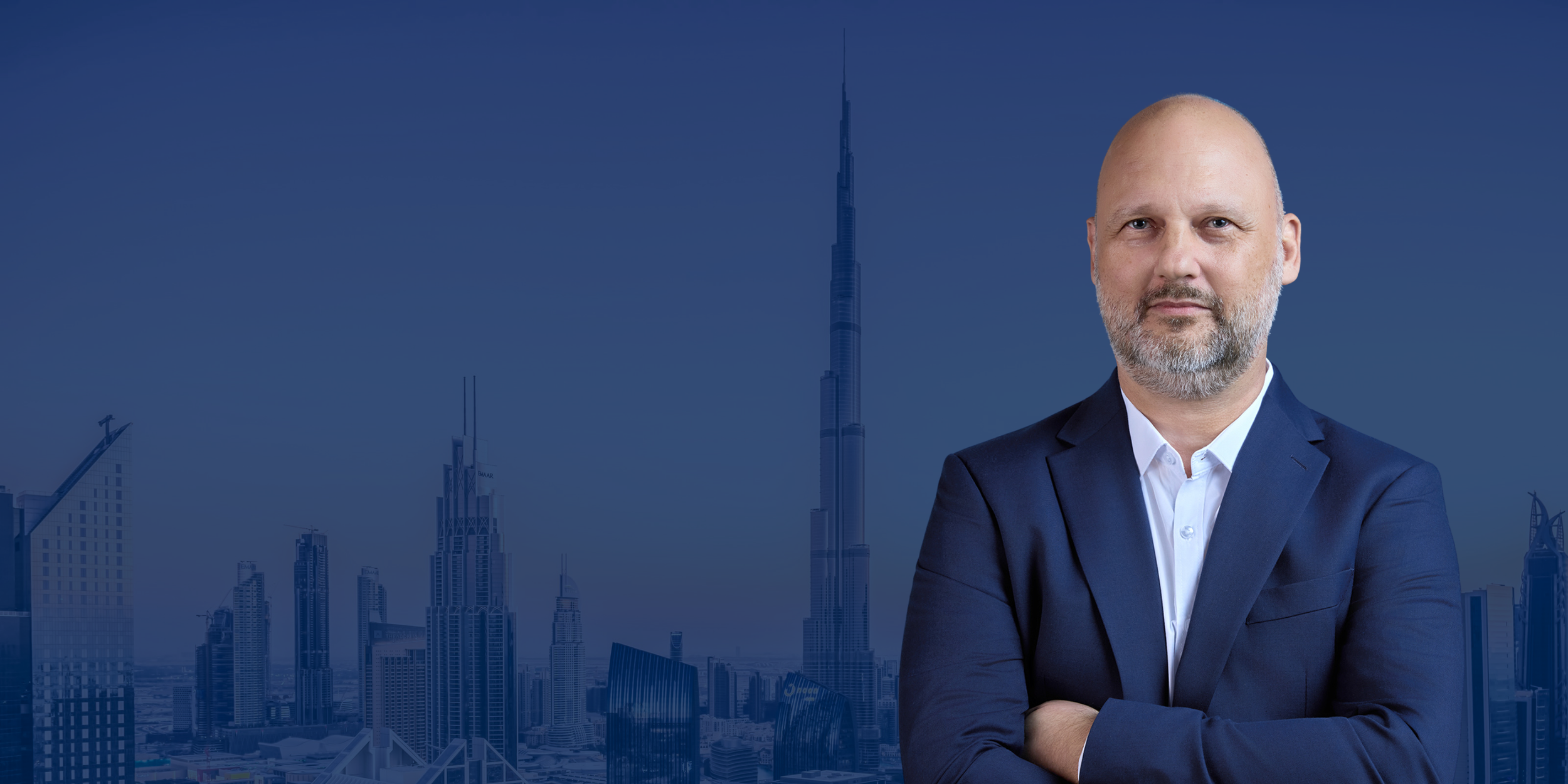 A man in a blue suit with a white shirt standing with arms crossed in front of the Dubai skyline, including the Burj Khalifa.