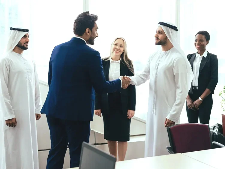 Two men, one in a dark suit and the other in traditional Middle Eastern attire, shake hands in a professional setting with three women watching, one of whom is smiling, all dressed in business attire.