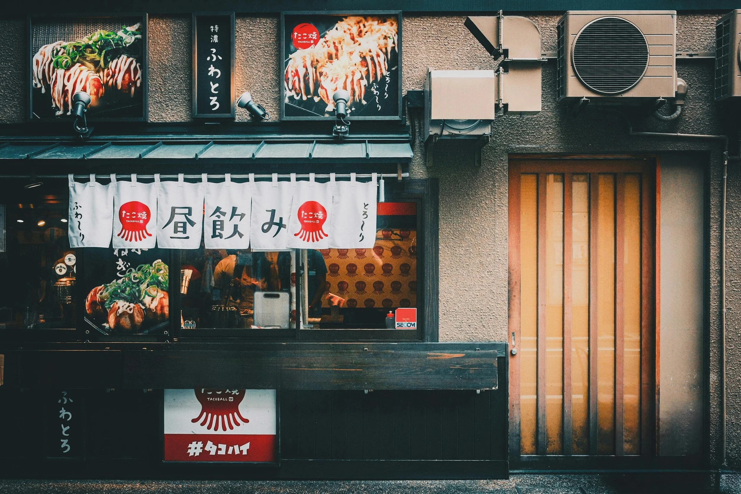 Esterno di un negozio di cibo giapponese con insegne e fotografie di piatti di takoyaki, con tende di stoffa bianche con scritte in giapponese e un'illustrazione di takoyaki rossi.