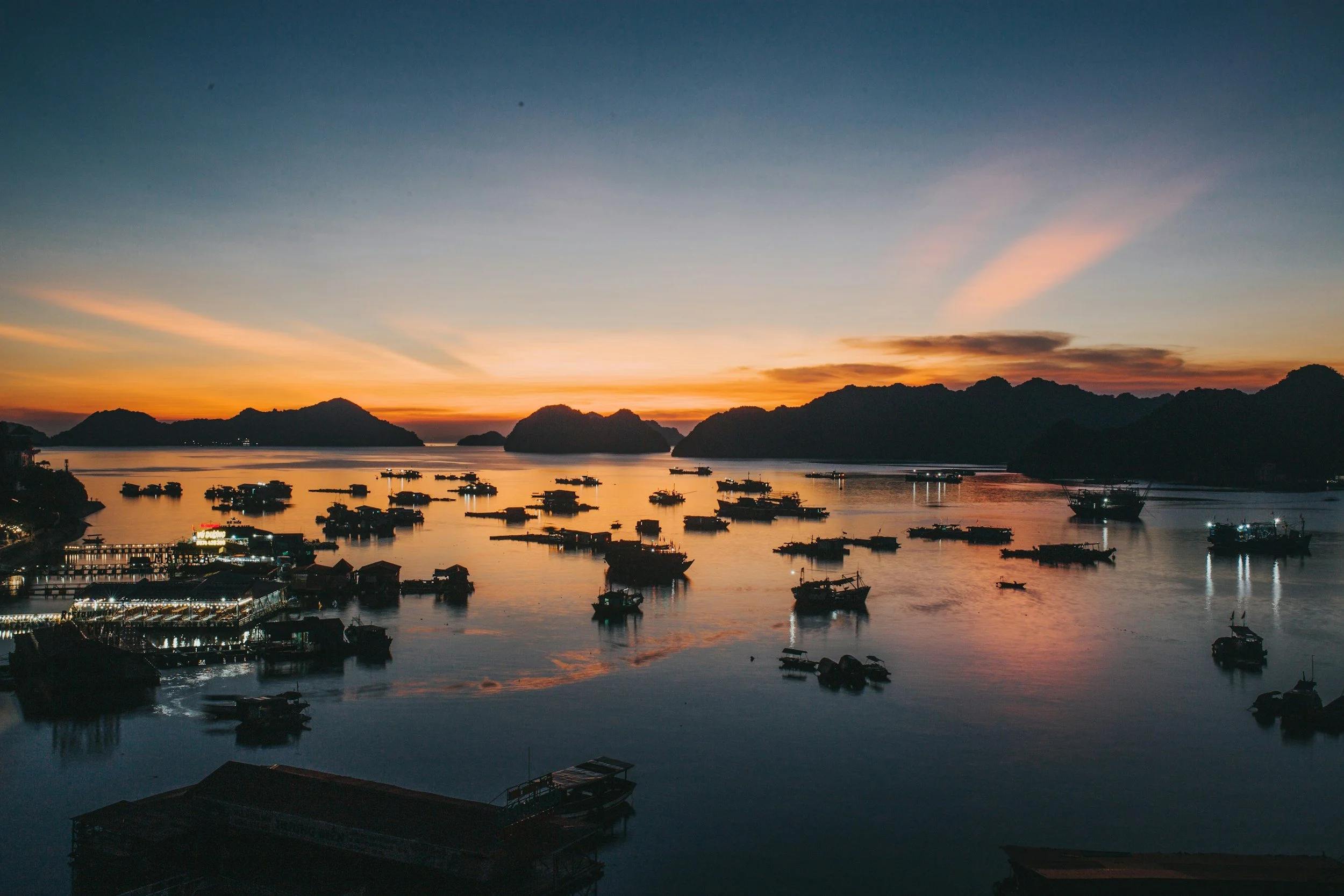 Paesaggio di un porto al tramonto con barche sul mare e montagne sullo sfondo.