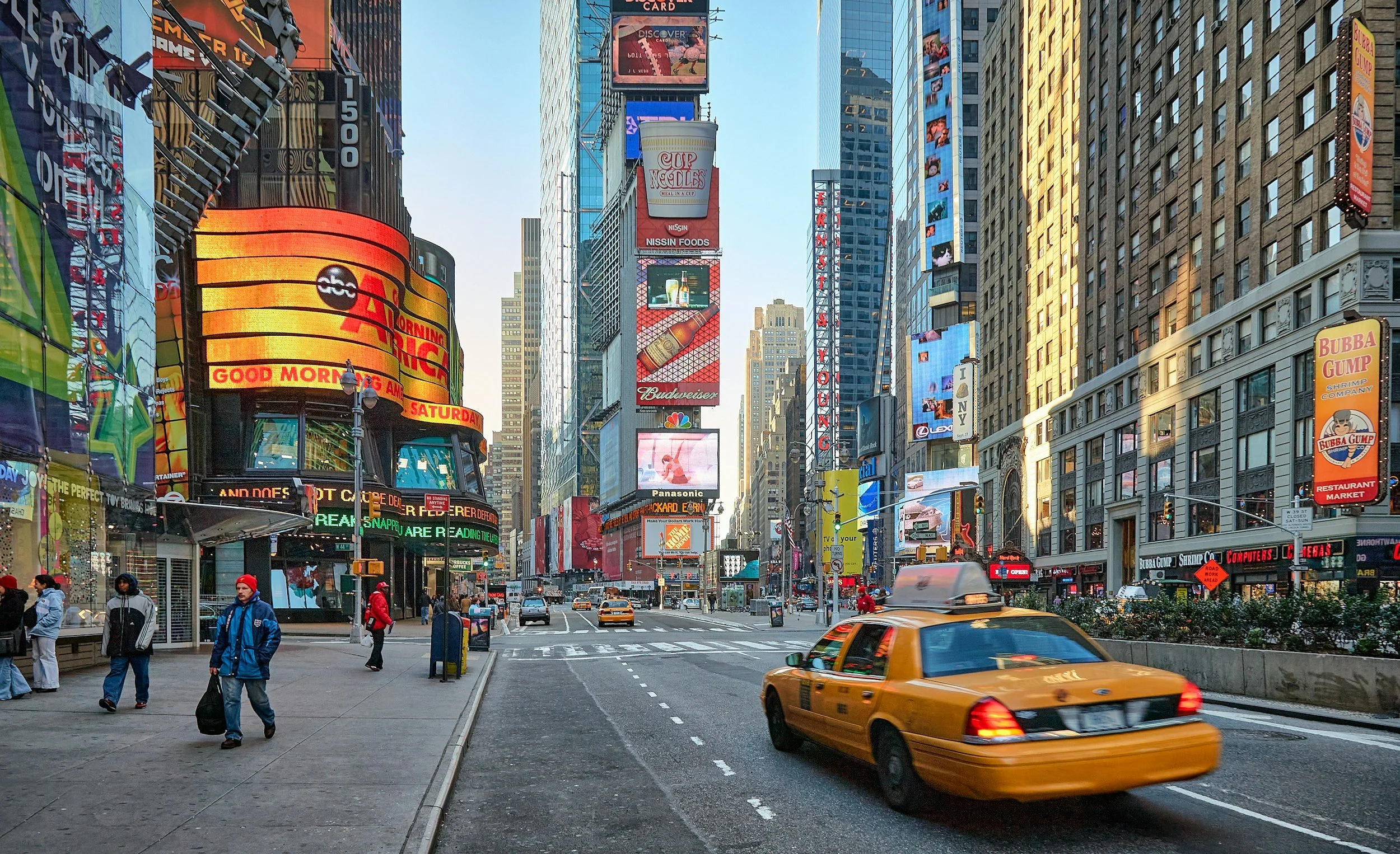 Una strada molto affollata a Times Square, New York, con grattacieli illuminati, cartelloni pubblicitari lampeggianti e un taxi giallo che passa sulla strada.