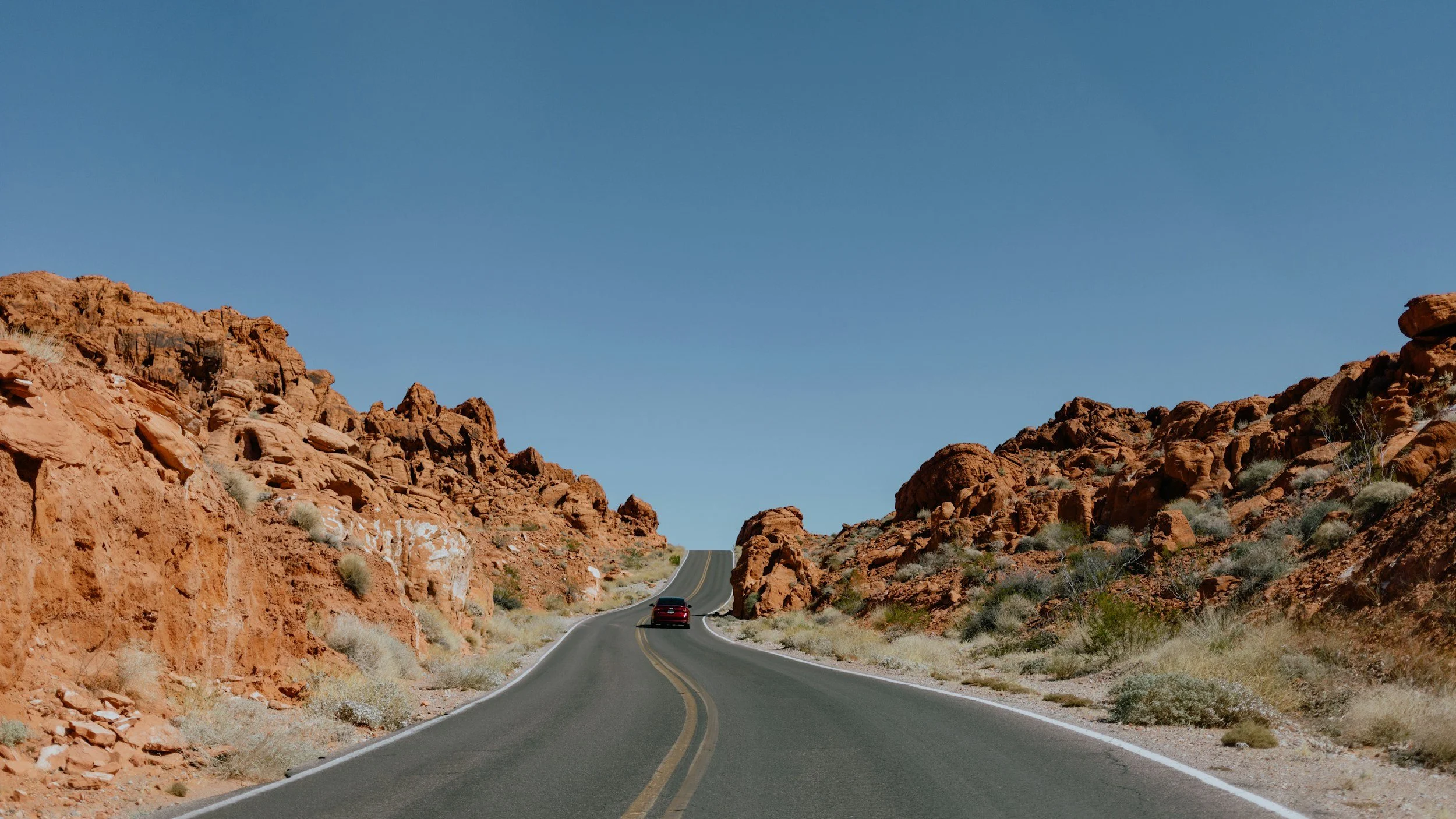 Strada che attraversa un paesaggio desertico con rocce rosse e sparse piante secche sotto un cielo chiaro azzurro.