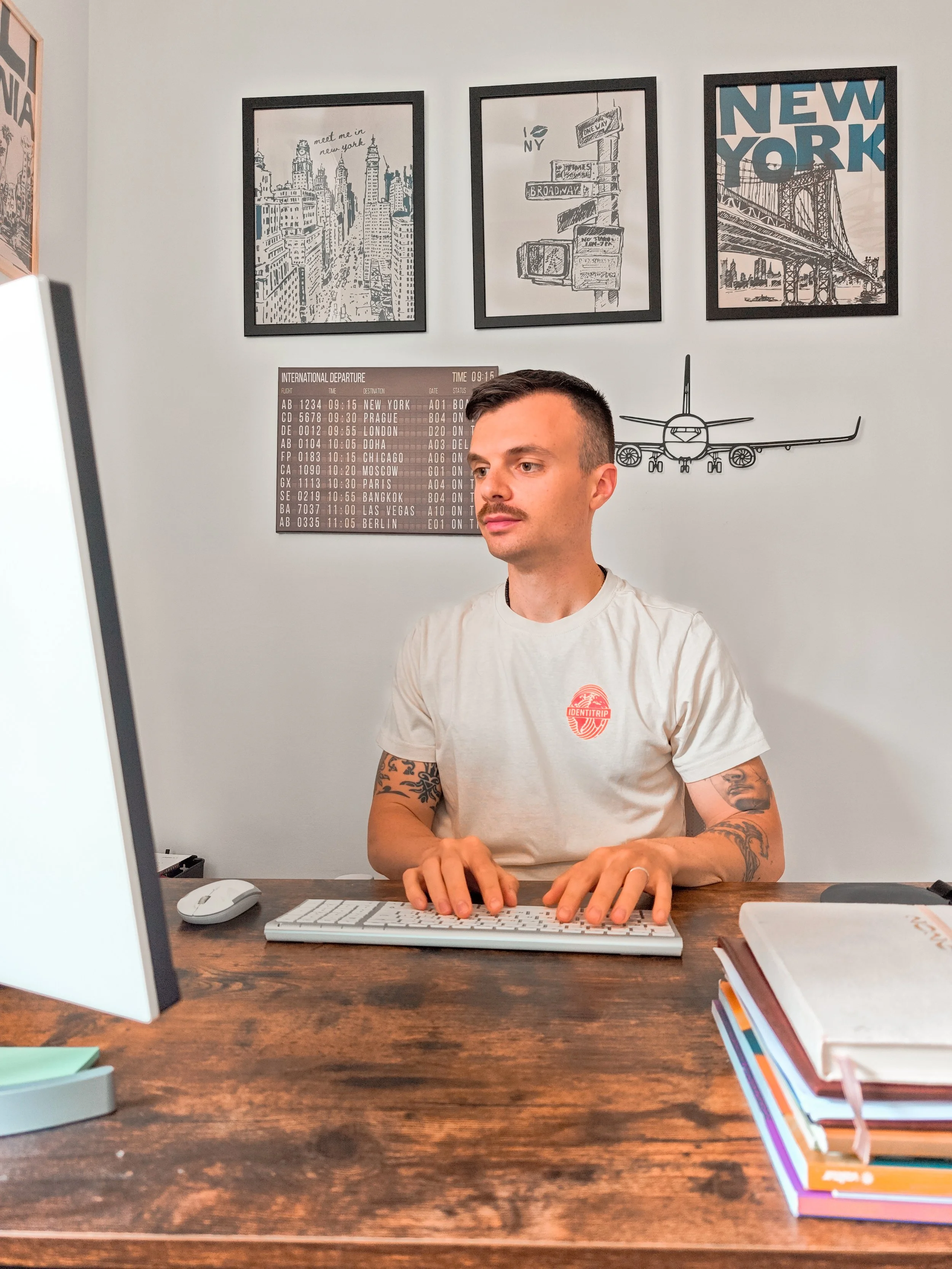 Uomo con tatuaggi alle braccia seduto alla scrivania che lavora al computer, con poster di New York e altri paesi sul muro e una pila di libri colorati a destra.