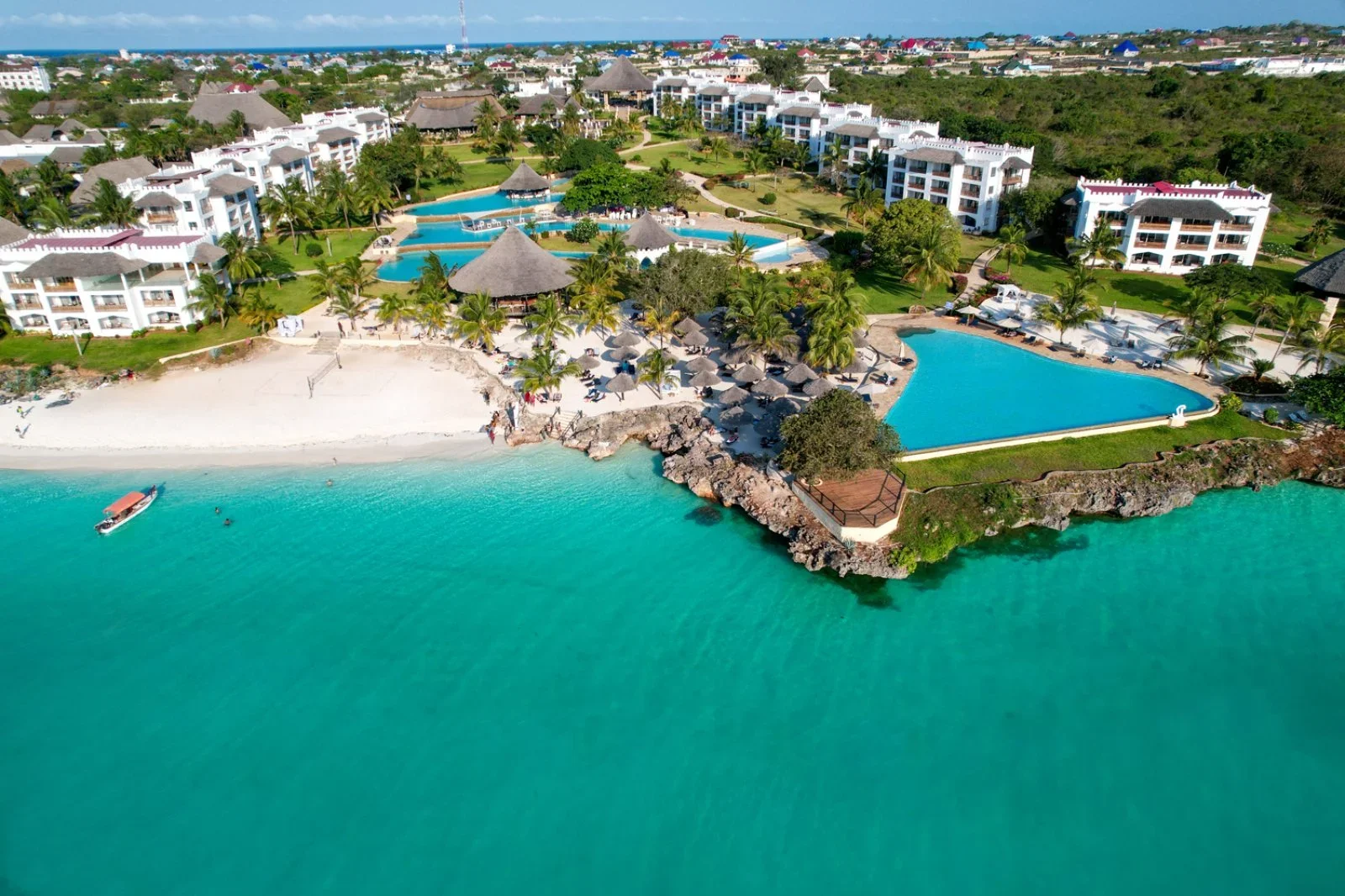 Vista aerea di un resort con piscine, spiaggia e mare turchese, con bungalow e case bianche tra palme e vegetazione