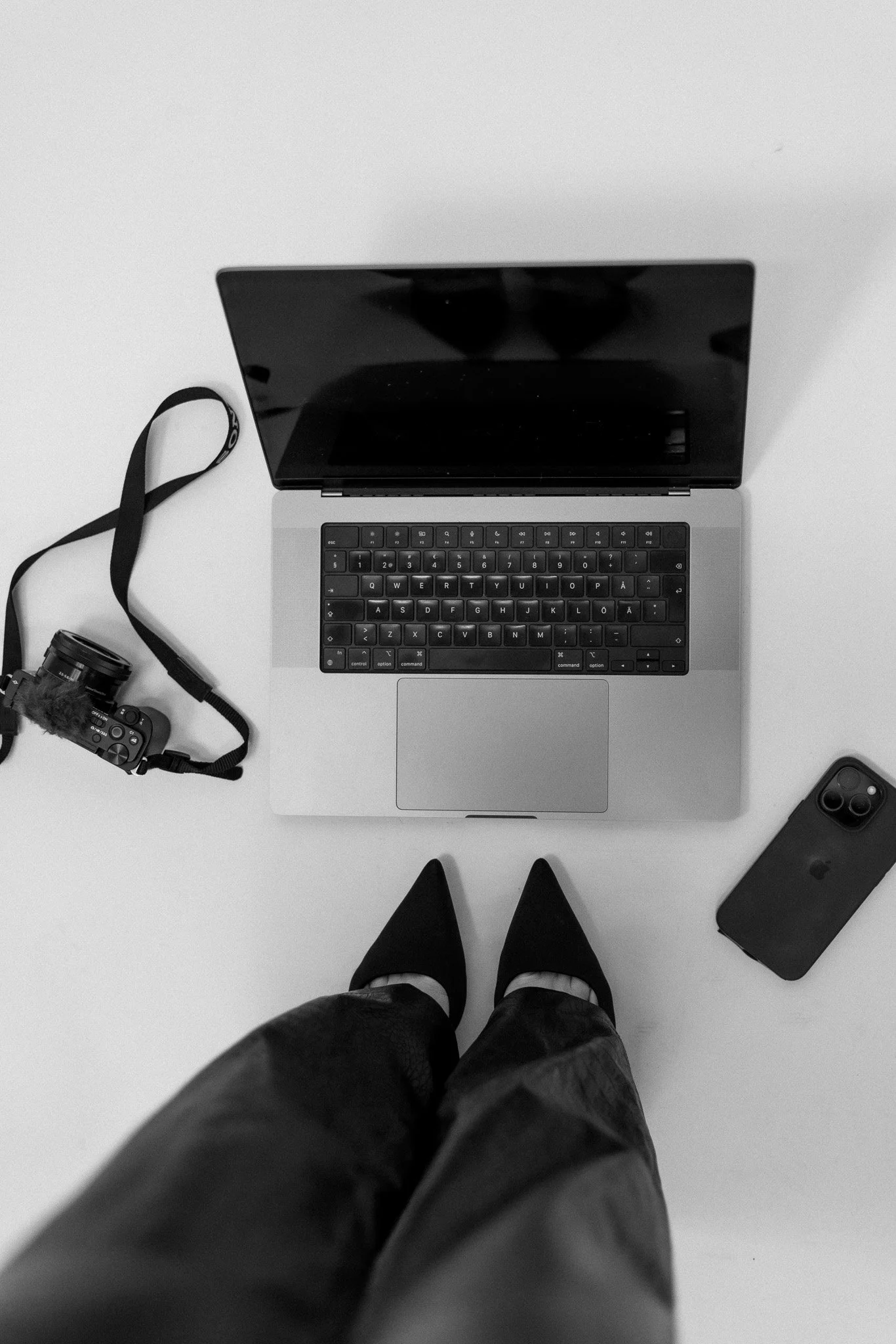 Henkilö katsoo alas nuoremman tietokoneen ja kameran päälle, jalat näkyvät ja hänellä on mustat korkokengät.